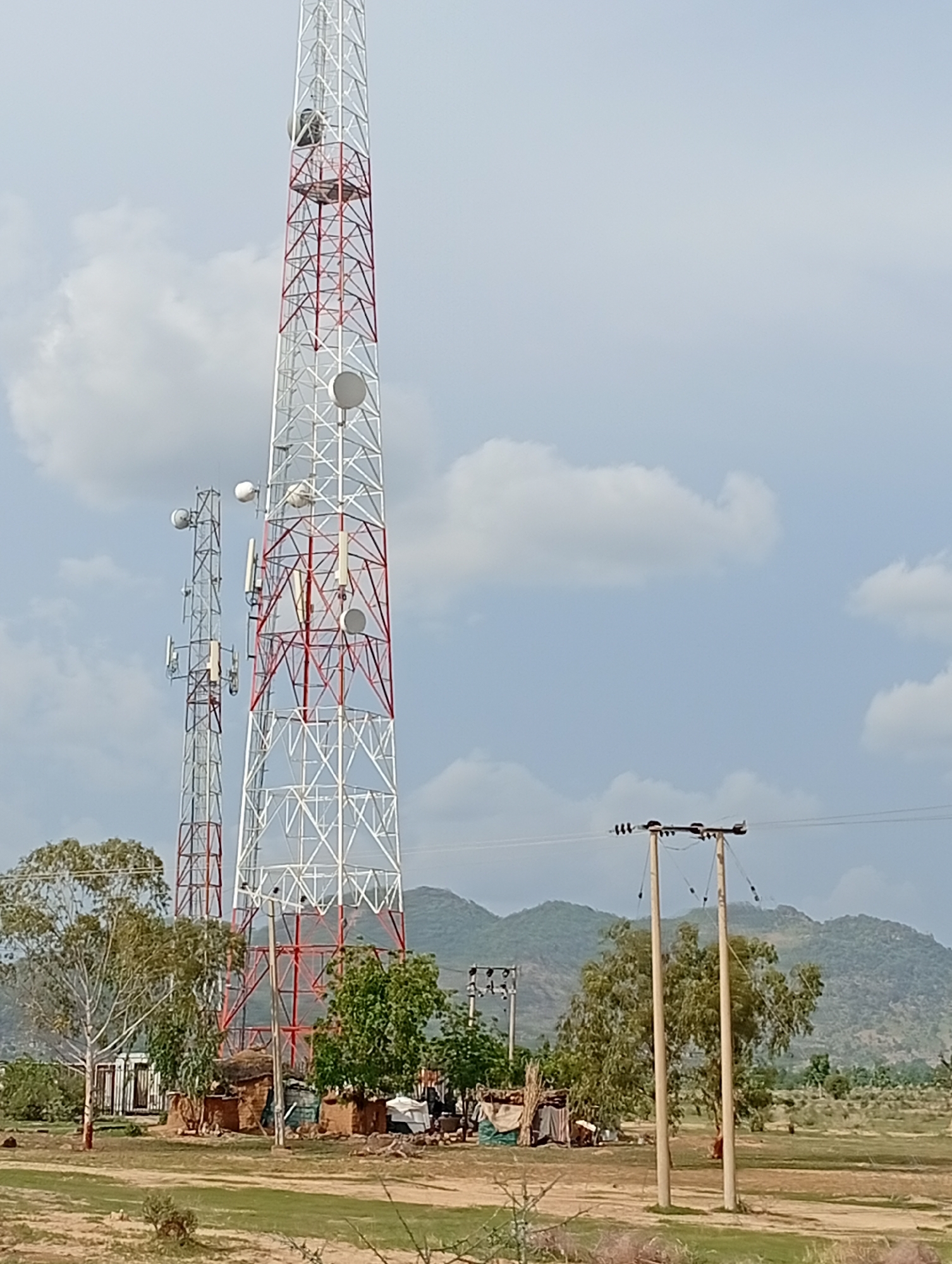 A wide-angle, outdoor, full shot captures a telecommunications tower and a smaller, adjacent tower in Shuwa, Nigeria. The main tower, painted white and red, is the dominant feature, with several circular dishes mounted on its structure. To its left, a second, thinner tower also supports communication equipment. In the foreground, scattered trees and small, rustic huts with thatched roofs are visible. In the background, rolling hills with sparse vegetation form a natural backdrop under a partly cloudy sky. The scene is characterized by a dry, open landscape, suggesting a rural or semi-rural setting within Shuwa.