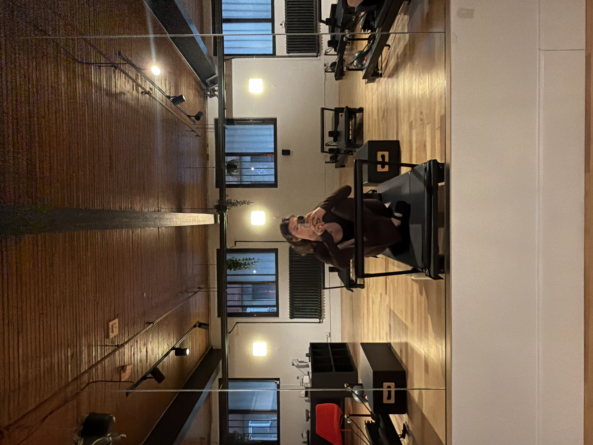 The image displays a modern fitness studio, likely a Pilates establishment, as seen through a large mirror selfie. A young woman, dressed in dark attire, is partially visible, holding a smartphone to capture her reflection. She appears to be focused on taking the photograph.
The studio features a spacious, well-lit interior. The flooring is made of light-colored wood, and the walls are a combination of clean white surfaces and a prominent, warm-toned wall covered in vertical wooden paneling. The ceiling reveals dark, exposed beams or ventilation ducts, contributing to an industrial-chic aesthetic.
Several black Pilates reformers with metallic frames are arranged throughout the room. One reformer in the foreground has a black box placed on it, featuring a distinctive white or light-colored 'C' cutout, possibly a brand logo or studio identifier. Other objects include black cubby-style storage units and a visible red upholstered seat. The studio is illuminated by square recessed lights on the ceiling and sleek, wall-mounted spotlight fixtures on the wooden paneling. Multiple rectangular windows with horizontal blinds or grates line the white walls, revealing a dark exterior, indicating it is likely evening or night. A large black radiator is situated beneath one of these windows, and a small potted plant adds a touch of nature near another window.
The setting suggests a contemporary and functional exercise space, consistent with urban fitness facilities found in locations like Montreal.