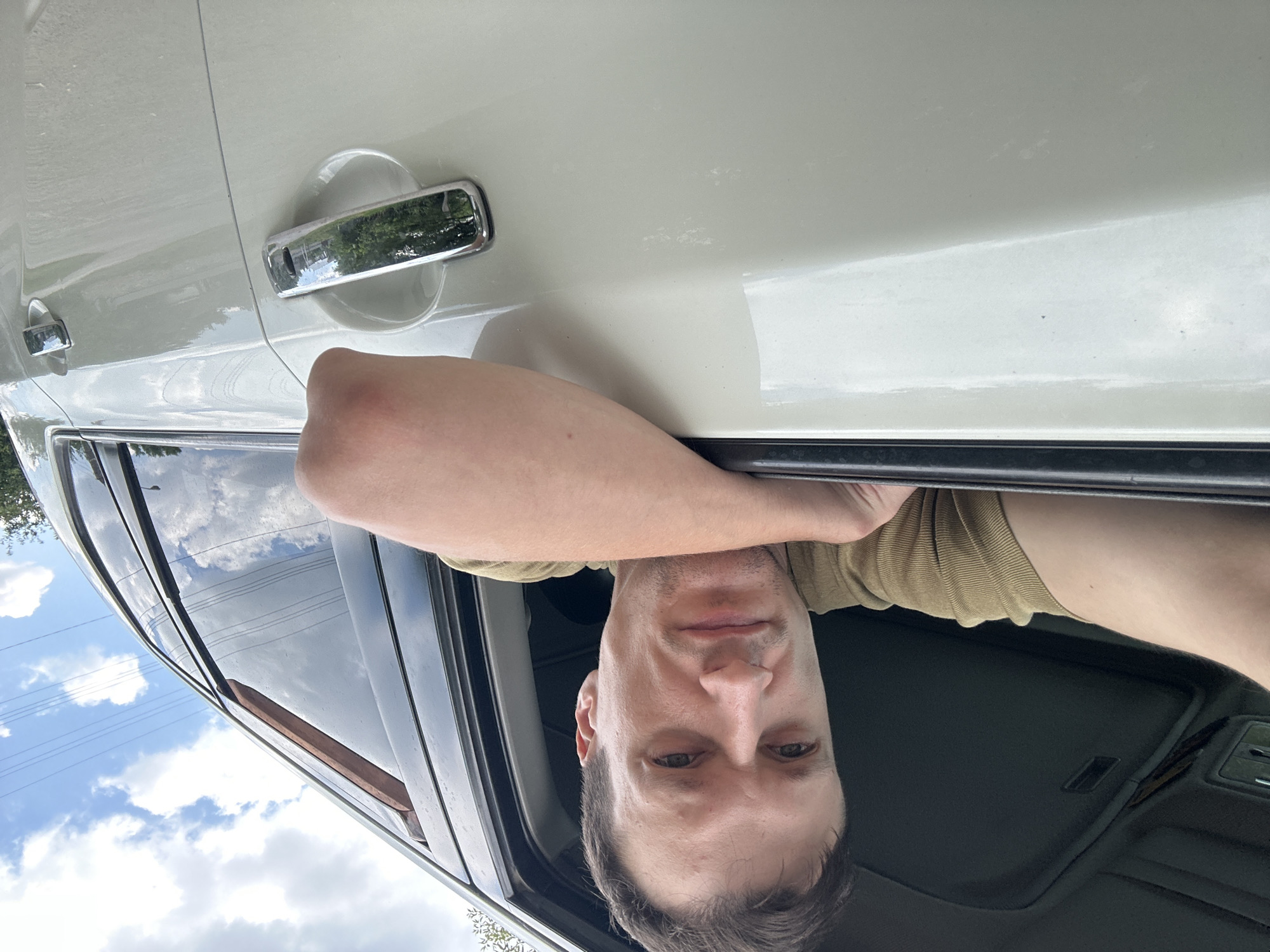 The image shows a man leaning out of a white SUV. He is wearing a khaki shirt and has his left arm resting on the window frame. The car appears to be parked outdoors, with a blue sky and clouds visible in the background. The man is looking directly at the camera with a neutral expression. The location is identified as Makiv, Ukraine, though no specific landmarks or urban features are visible in the image to corroborate this. The scene is a close-up shot, focusing primarily on the man and the side of the vehicle.