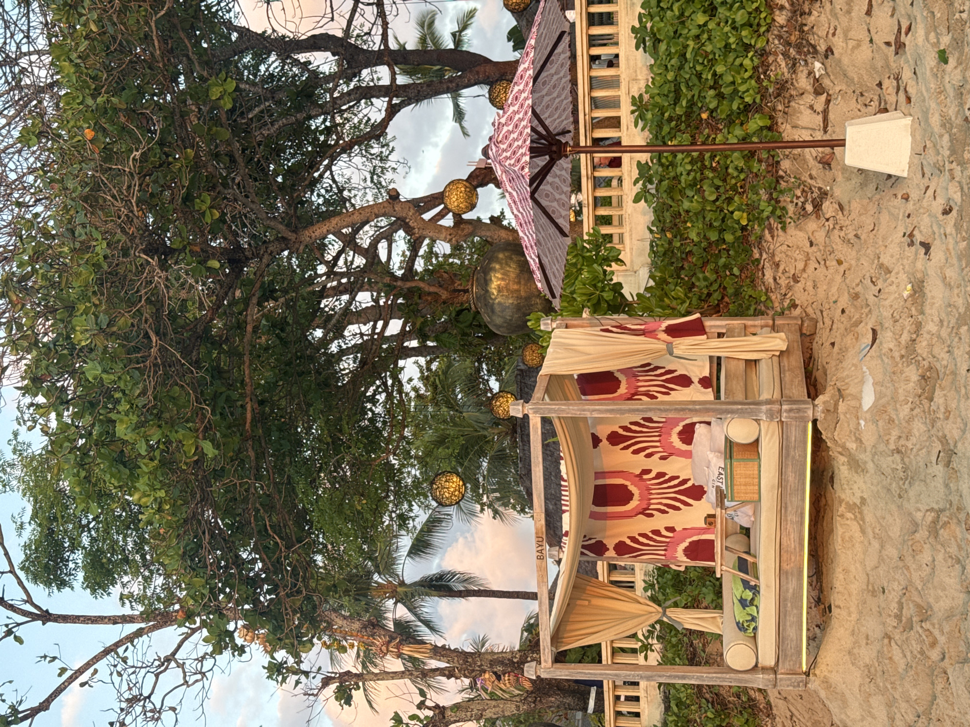 The image shows a beach scene in Kuta Selatan, Indonesia. A wooden cabana with patterned fabric and cushions sits on the sand. A large tree with bare branches and green leaves provides shade. Decorative golden orbs hang from the branches. An umbrella with a similar pattern is partially visible behind the cabana. The background includes a building structure and green vegetation. The sand is light beige, and the sky is visible through the trees.