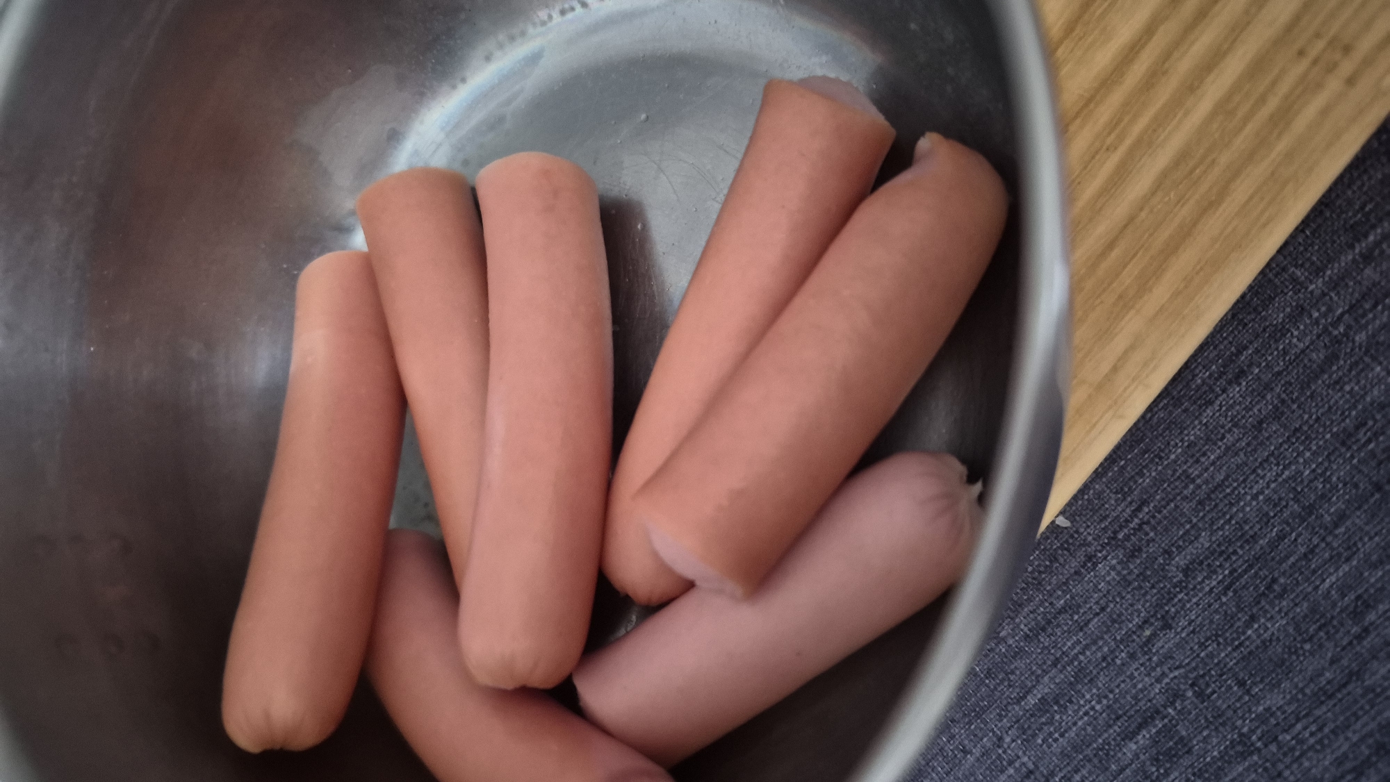 The image shows a stainless steel bowl containing several uncooked hot dogs. The hot dogs are a light pinkish-orange color and are arranged in a cluster within the bowl. The bowl is placed on a surface that appears to be a combination of dark blue fabric and a light brown wooden surface. There are no people or any other identifiable objects in the image. The image does not provide any information about the location in the city of Krauja, Latvia.