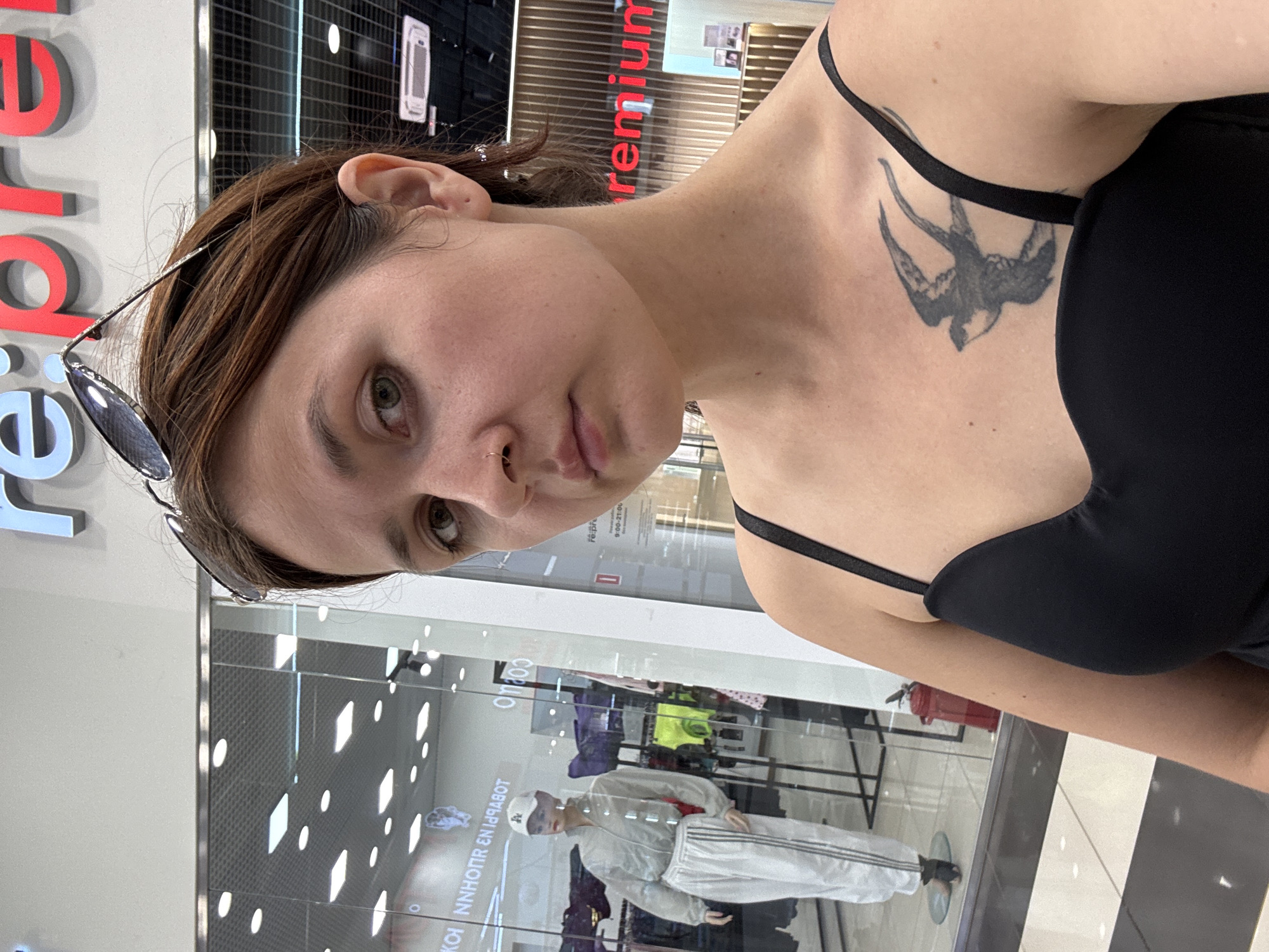 A young woman with auburn hair and a bird tattoo on her chest takes a selfie in a shopping mall. She is wearing a black tank top and has sunglasses on top of her head. In the background, a mannequin dressed in a light-colored tracksuit is visible through a store window. The store's name, "re:premium," is visible in large red lettering on the wall behind her. The flooring consists of black and white tiles. The scene is likely inside the "Ecdotic" shopping center in Naberezhnye Chelny, Russia, given the visual cues of retail storefronts and mall aesthetics.