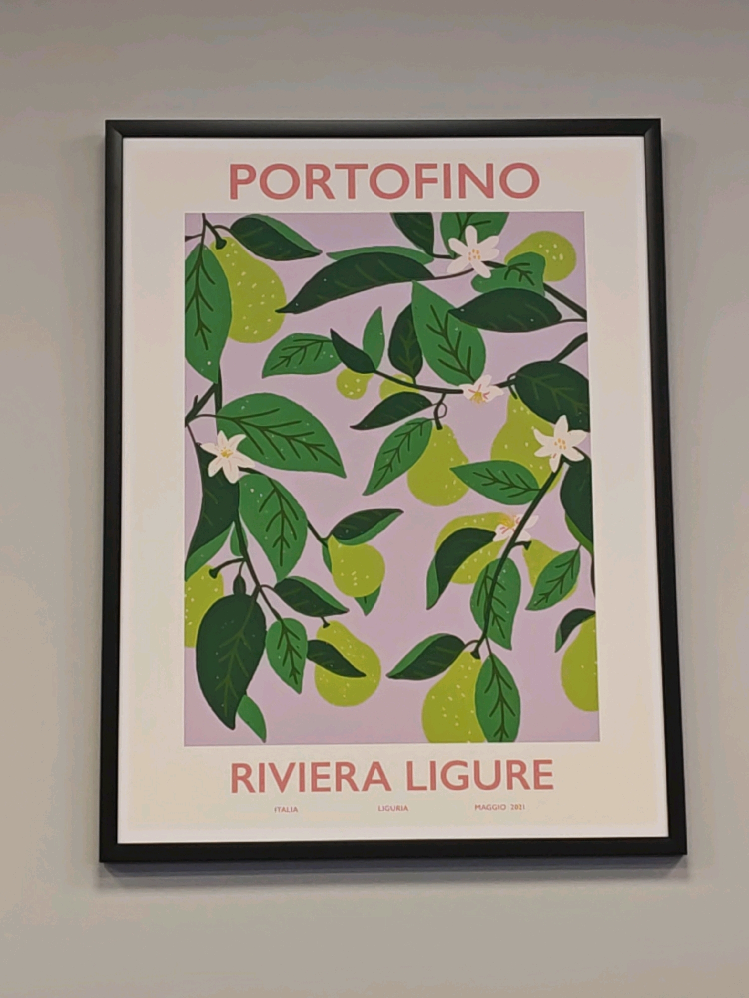 The image shows a framed poster hanging on a plain, light-colored wall. The poster features a stylized illustration of pears and leaves against a pale purple background. At the top of the poster, in large, pink sans-serif letters, is the word "PORTOFINO." Below the illustration, in smaller pink letters, is "RIVIERA LIGURE," with "ITALIA," "LIGURIA," and "MAGGIO 2021" written below it in even smaller font. The frame is black and thin. The wall appears to be an interior wall with no visible windows or other decorative elements. The lighting is even, suggesting an indoor setting. The image is taken from a slightly angled perspective, with the top edge of the poster angled slightly away from the viewer. There are no people or animals visible in the image. The location is not Saratov, Russia.