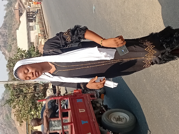 A woman wearing a black dress with gold detailing and a white headscarf stands on the side of a road. She's holding a phone and some white fabric. Behind her, a red three-wheeled vehicle is visible, and in the background are buildings and a hill. The scene is bathed in bright sunlight, suggesting a clear day.