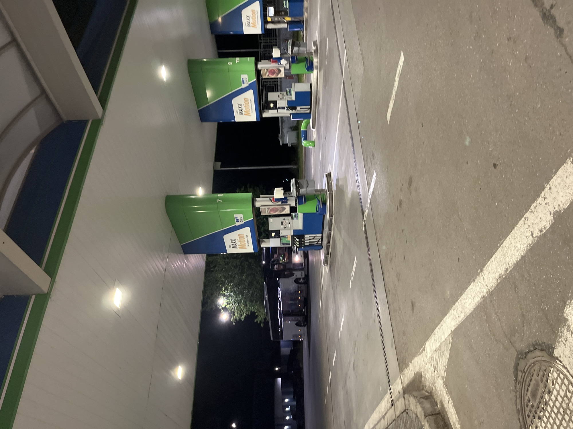 The image depicts a Maxx Motion gas station in Popovitsa, Bulgaria at night. Several gas pumps are visible under green and blue canopies, marked with numbers and branding. A large bus is parked near the pumps, casting elongated reflections. The surrounding area shows a paved lot with painted white lane markings. The environment suggests a typical roadside gas station setup along a street with streetlights. A manhole cover is on the edge of the road.