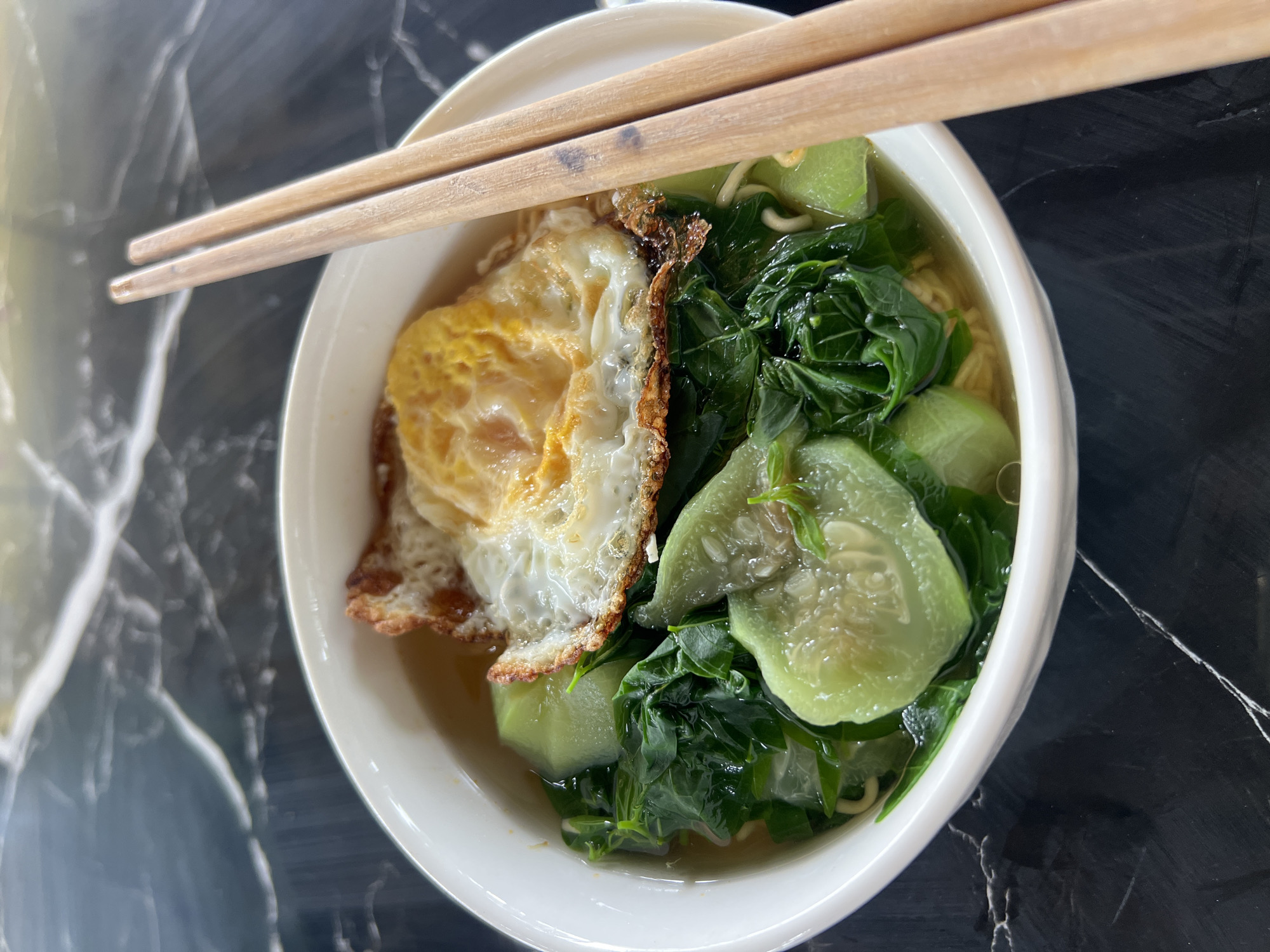A close-up, high-angle shot shows a white ceramic bowl filled with noodle soup, topped with a fried egg, leafy green vegetables, and light green sliced vegetables. Two wooden chopsticks rest on the right side of the bowl, poised over the food. The bowl sits on a dark, polished surface with a marble-like pattern. The scene appears to be an indoor setting, possibly a dining area, in Thái Sơn Commune, Vietnam.