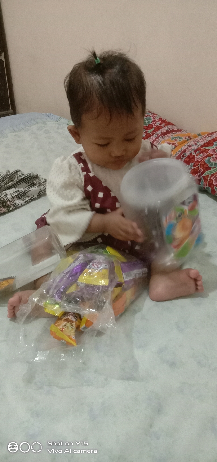 A single infant is seated barefoot on a light-colored, textured mattress in an indoor domestic setting, presumed to be in Ranugedang, Indonesia. The infant, with dark hair tied in a small top-knot, wears a white textured long-sleeved top and a maroon pinafore with white square patterns. The infant is looking downwards, with the right hand holding a transparent plastic jar containing blurry contents and featuring a colorful label. To the infant's left, a clear plastic container with its lid open is visible. In front of the infant, a clear plastic bag holds multiple individually wrapped snacks in yellow, purple, and orange packaging. A red and white patterned pillow is behind the infant's right side, and a folded patterned blanket is on the left. The background wall is a plain light pink hue.