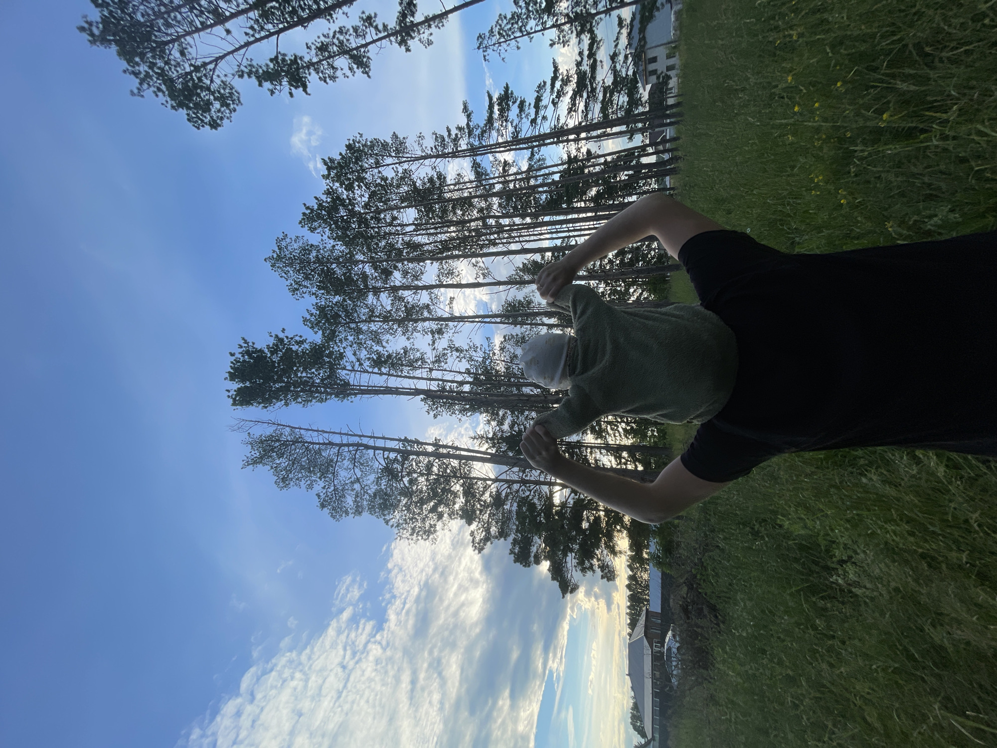 The media file shows a person lying on their back in a grassy field, looking up at the sky. They are wearing a green shirt and black shorts, and a white cap. The person's arms are raised, and they appear to be holding something in their hands, though it is not clearly visible.
The scene is set in a rural area with tall pine trees in the background, and a few houses visible in the distance. The sky is blue with scattered white clouds, suggesting it is daytime, likely late afternoon given the warm light. The overall mood seems peaceful and relaxed.
The location is identified as Подшивалово, Russia. There is no visible text in the image.