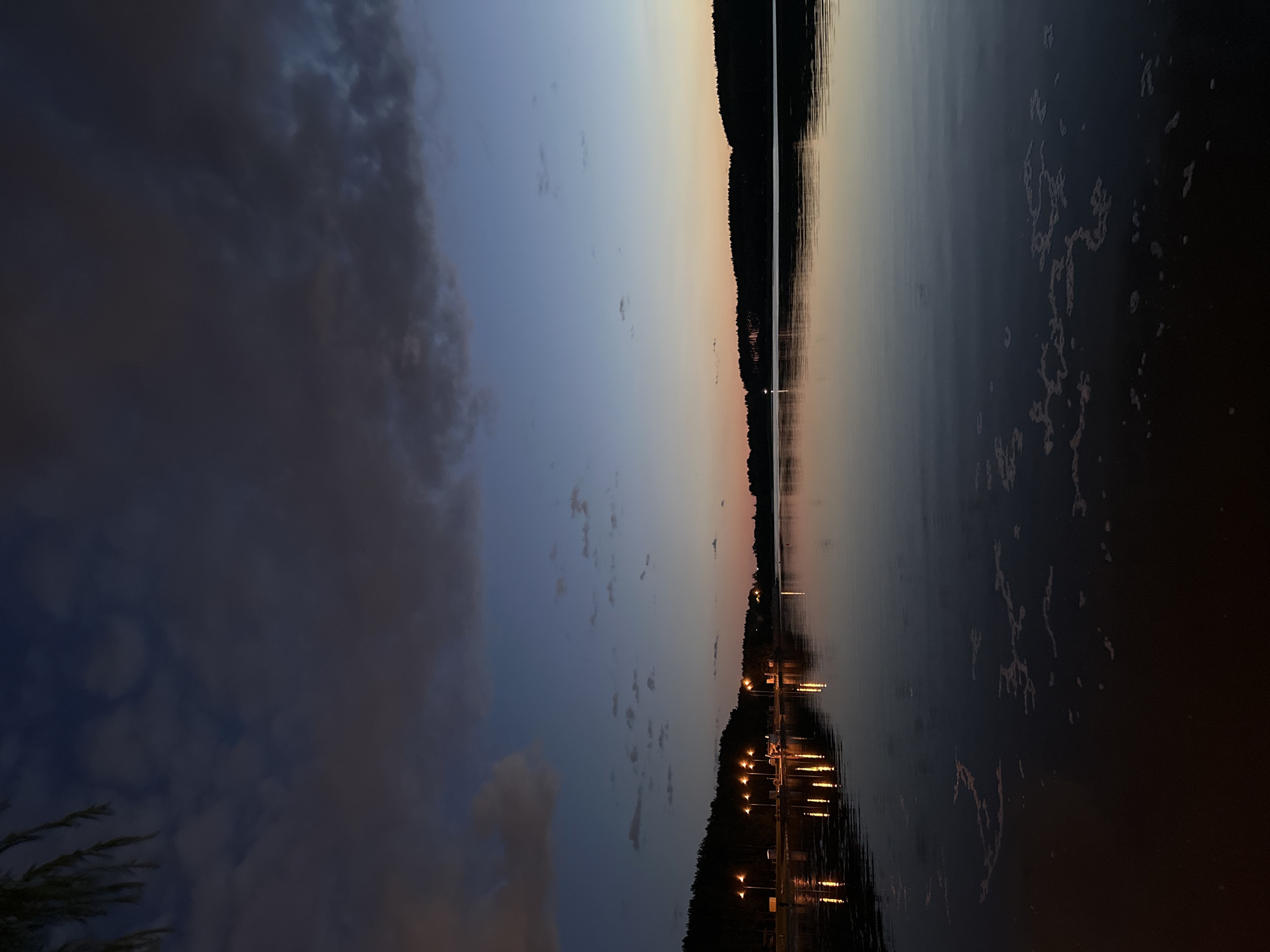 The image depicts a serene lakeside scene at dusk, likely in Minsk, Belarus, given the provided context. The sky transitions from a deep blue overhead to softer hues of orange and pink near the horizon. Dark, silhouetted trees line the distant shore. On the left side of the image, a row of illuminated streetlights casts reflections onto the calm water. The water's surface shows subtle ripples and patches of foamy debris. The overall atmosphere is tranquil and peaceful, evoking the quiet beauty of a lakeside evening.