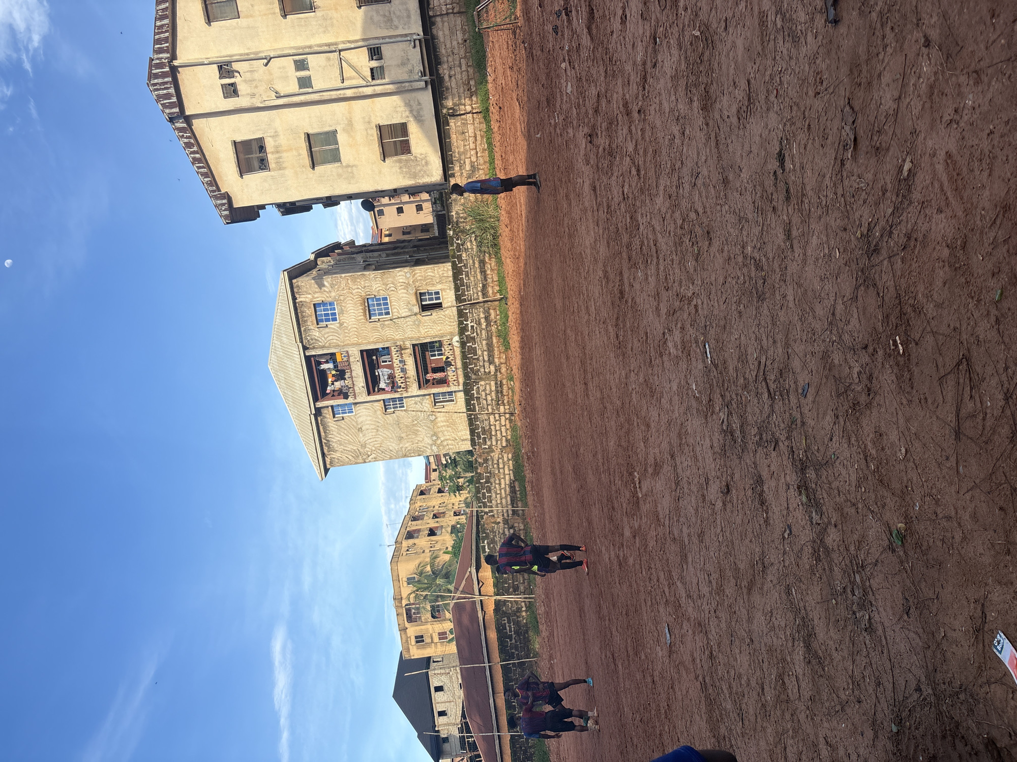 The image depicts a natural dirt soccer field in Onitsha, Nigeria, with several individuals engaged in a game. The field is rough, uneven, and a reddish-brown color, indicative of local soil. In the background, residential buildings with multiple stories are visible. One building features balconies adorned with laundry, suggesting active habitation. The sky is a clear blue with a few wispy clouds and the moon visible. The scene appears to be set in the late afternoon or early evening, as indicated by the lighting. The overall impression is of a community gathering for recreation in an urban environment.