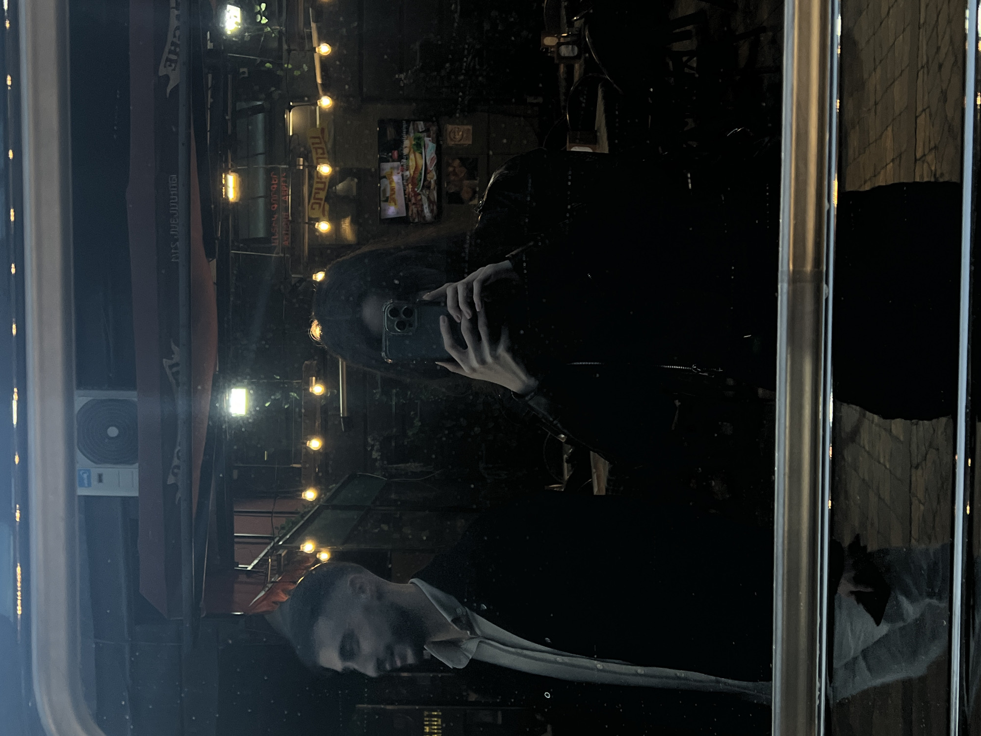 A nighttime reflection scene in a window or glass surface shows two individuals and an external environment. One individual is taking a picture with a smartphone, holding the device up at face level. The second person is positioned beneath, seen in profile, wearing a light-colored collared shirt and potentially a darker suit jacket. The reflected background consists of a cityscape with buildings, some visible signage in a non-English script, and decorative string lights. There's also a reflected image of what appears to be a menu board or a display with food items. An air conditioning unit is visible on the left, near the top of the image, outside the glass. The specific location in the city of Unknown, Unknown cannot be determined from the available information.