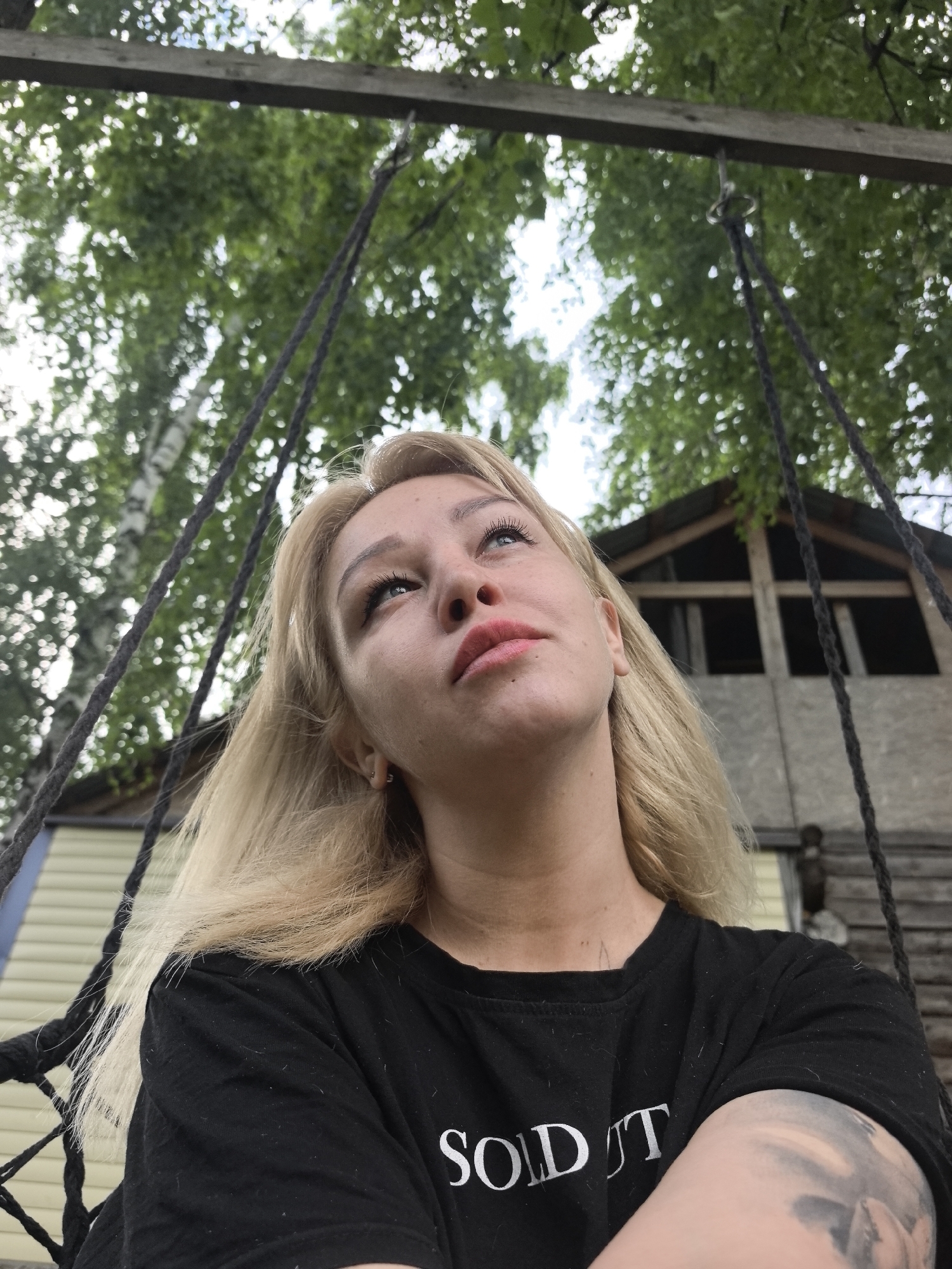 A young woman with blonde hair and blue eyes is sitting on a swing, looking upwards. She is wearing a black t-shirt with white lettering that appears to read "SOLD IT". Her right arm is crossed over her chest and displays a tattoo. The background consists of lush green trees and a wooden structure that resembles a treehouse or cabin. The setting appears to be outdoors in a natural environment, possibly a backyard or park. There is no indication of the specific location within the city of Noviy, Russia.
