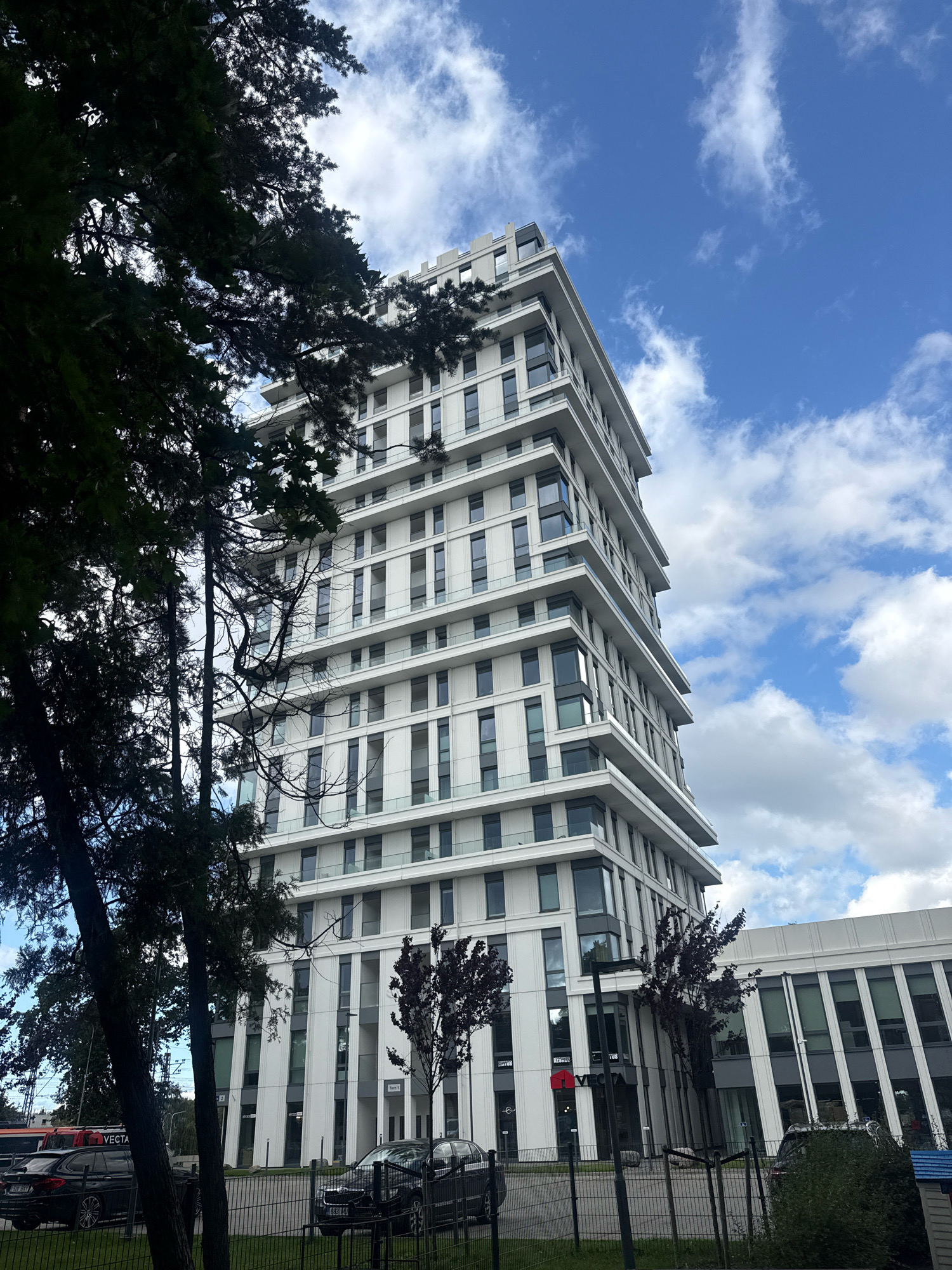 This is a low-angle, full-shot photograph of a modern, multi-story building with a white facade and dark-tinted windows, set against a bright blue sky with scattered white clouds. The building has a stepped design, with each successive floor recessed slightly from the one below. The facade is characterized by vertical white bands and horizontal gray balconies. In the foreground, to the left, are the dark green, leafy branches of a tall tree, partially obscuring the view of the building. To the right of the main building, there is a lower, horizontal section of the complex, also with a white facade and rows of dark-tinted windows. A black fence runs along the bottom of the image, with a patch of green grass behind it. Several black cars are parked behind the fence. The building is identified by a red VECTA logo on the lower, horizontal section. This building is located in Tallinn, Estonia.