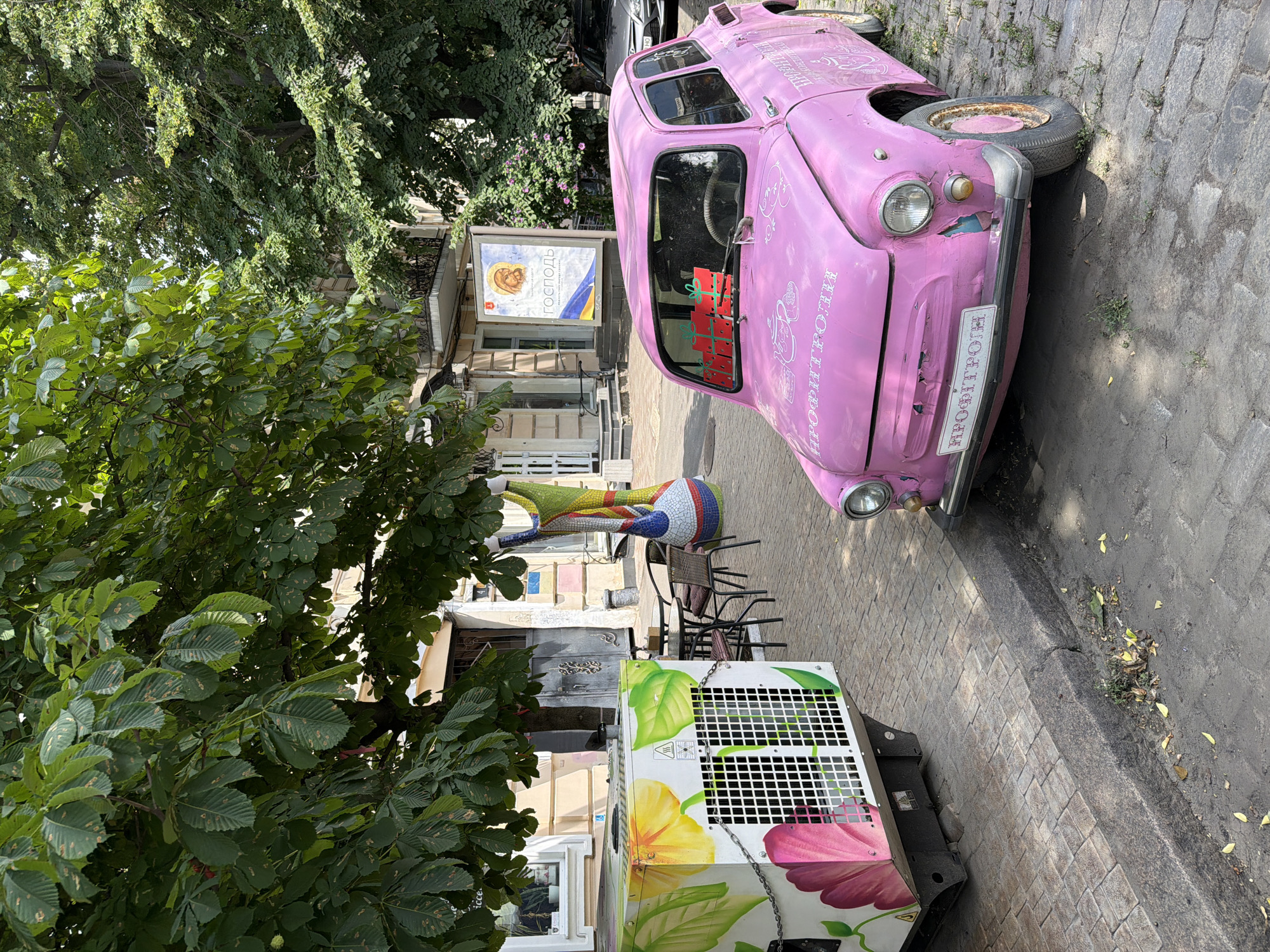 The image depicts a street scene in Odesa, Ukraine, with a vibrant pink vintage car parked on a cobblestone street. The car, possibly a ZAZ or similar Soviet-era model, is adorned with white line drawings, including teacups and the word "ПРОФІТРОЛІ" (Profitrolli), suggesting a bakery or confectionery theme. Decorative red gift boxes are attached to the car's windshield.
To the left of the car, a large, white, rectangular air conditioning unit or similar piece of equipment is decorated with brightly colored floral patterns, featuring large pink and yellow flowers. Behind the AC unit, a small outdoor seating area with metal tables and chairs is visible, suggesting a cafe or restaurant.
In the background, buildings line the street, one with a sign featuring an image of Christ and text in Cyrillic, and a banner with the Ukrainian flag. A tall, colorful, mosaic-like sculpture stands near the outdoor seating area. Lush green trees with large leaves overhang the scene, providing shade. The overall environment appears to be a charming, possibly pedestrian-friendly, street in Odesa, characterized by its artistic decorations and vintage elements.