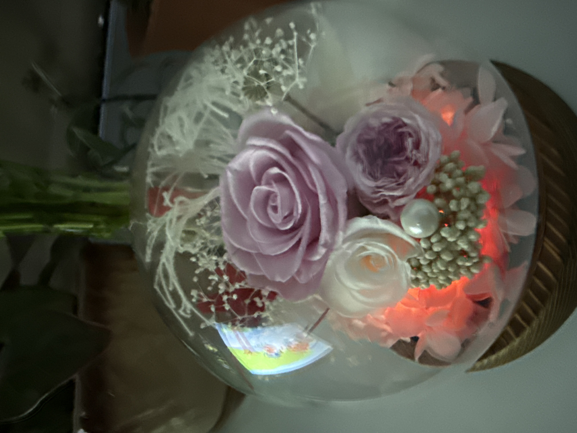 A preserved flower arrangement sits in a clear glass dome, illuminated by a soft red light from within. The arrangement features a large, light purple rose at the top, with smaller white and pink roses below. Tiny white filler flowers and red accents are scattered throughout. A single pearl is positioned in the center of a cluster of small, light-colored beads. The dome is supported by a wooden base. The background is dark and out of focus, suggesting an indoor setting. There is no indication of the city of Cheboksary, Russia.