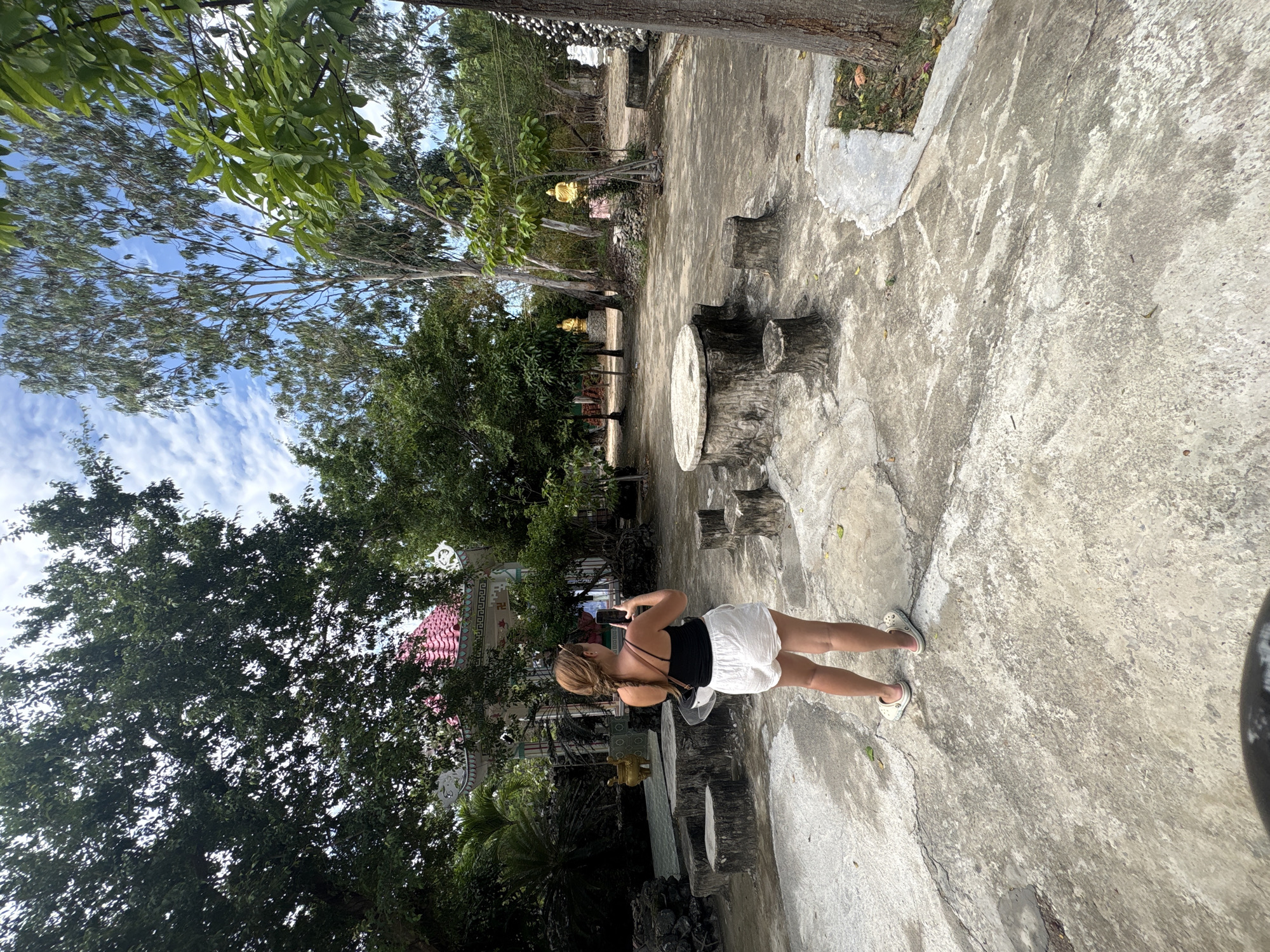 This image captures a bright, sunny daytime scene in what appears to be a temple or garden complex in Cam Ranh, Vietnam. The main subject is a person, seen from behind, standing on a concrete path. They are wearing white shorts, a black sleeveless top, and white Crocs, with their hair in a braid and sunglasses on their head. The person is holding a smartphone up, seemingly taking a photo or video of the area in front of them.
The setting is characterized by a mix of natural elements and man-made structures. The ground is largely concrete, showing signs of wear and some cracks, with occasional patches of soil and grass. To the right of the person, there's a stone or concrete table and two stools, carved to resemble tree stumps.
The background is dominated by a variety of green trees and lush foliage under a clear blue sky dotted with scattered white clouds. Amidst the trees, traditional Asian architecture is visible, including a building with a prominent red-tiled roof and white ornate decorative elements. A swastika symbol, commonly found in Buddhist temples, is discernible on this structure. Further into the mid-ground, partially obscured by greenery, at least two golden Buddha statues can be seen, reinforcing the impression of a religious site. The overall mood is peaceful and serene. No explicit text is visible in the image.