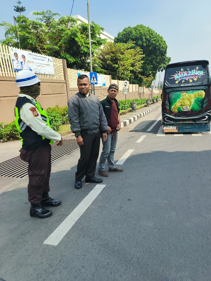 The image depicts a scene in Ngamprah, Indonesia (latitude: -6.866, longitude: 107.515). A uniformed traffic officer, wearing a green vest and a white helmet, stands on the left side of the road, facing the right. He appears to be talking to two men who are standing on the road. The man on the left is dressed in a grey jacket and dark trousers. The man on the right is wearing a red shirt and blue jeans. A brightly colored bus, with the word "AMAZING" painted on its side, has just parked on the right side of the road, facing away from the camera. The road is paved and has a white dividing line in the middle. The background is a cityscape with a row of trees.