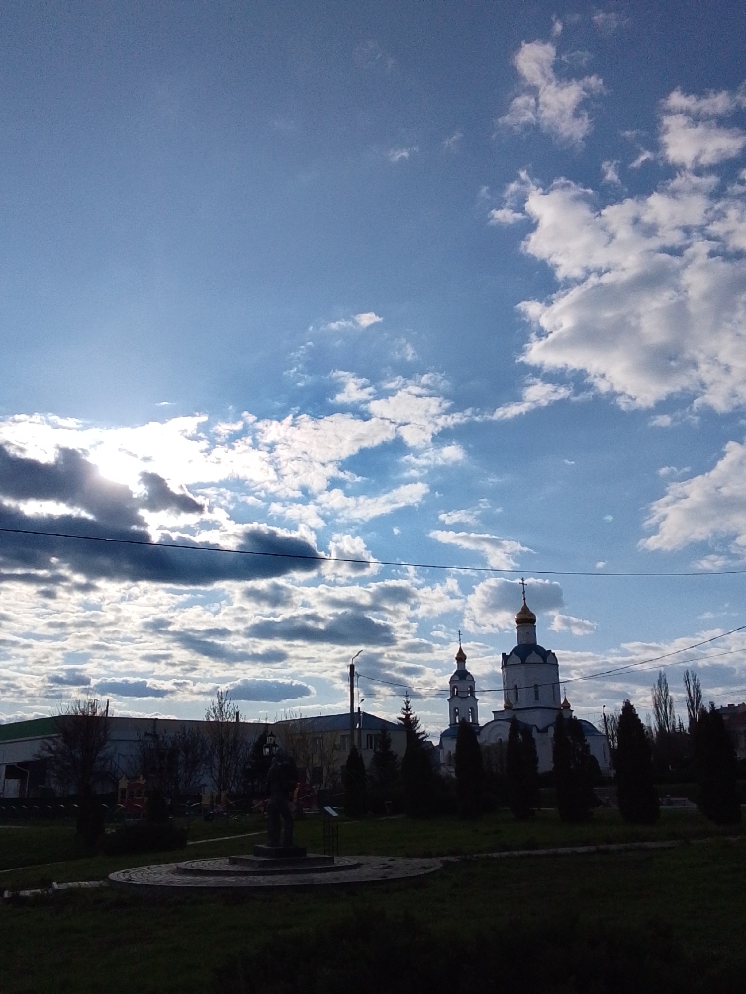 The image shows an outdoor scene in Khlevnoye, Russia, under a partially cloudy sky. A church with a white facade and gold domes is visible in the midground. In the foreground is a dark park with a statue on a pedestal. The area in front of the statue is a paved, circular platform. Several coniferous trees and bare deciduous trees are scattered throughout the park. In the background, a low-rise building with a green roof and adjacent building are visible. Power lines are strung across the sky.