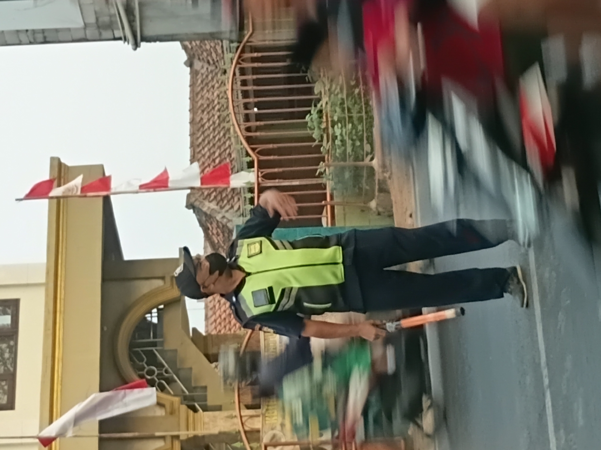 A man wearing a dark uniform, a bright lime green vest with reflective stripes, and a black baseball cap with a logo is standing on a street, holding an orange traffic baton. He is also wearing a black face mask. The man appears to be directing traffic. In the background, there are motorcycles in motion, indicating traffic is present. A building with an arched entrance and stairs is visible behind him, with Indonesian flags flying. The scene appears to be in an urban environment in Bekasi, Indonesia, given the context of the flags and the urban setting. The overall lighting suggests it is either early morning or late afternoon.