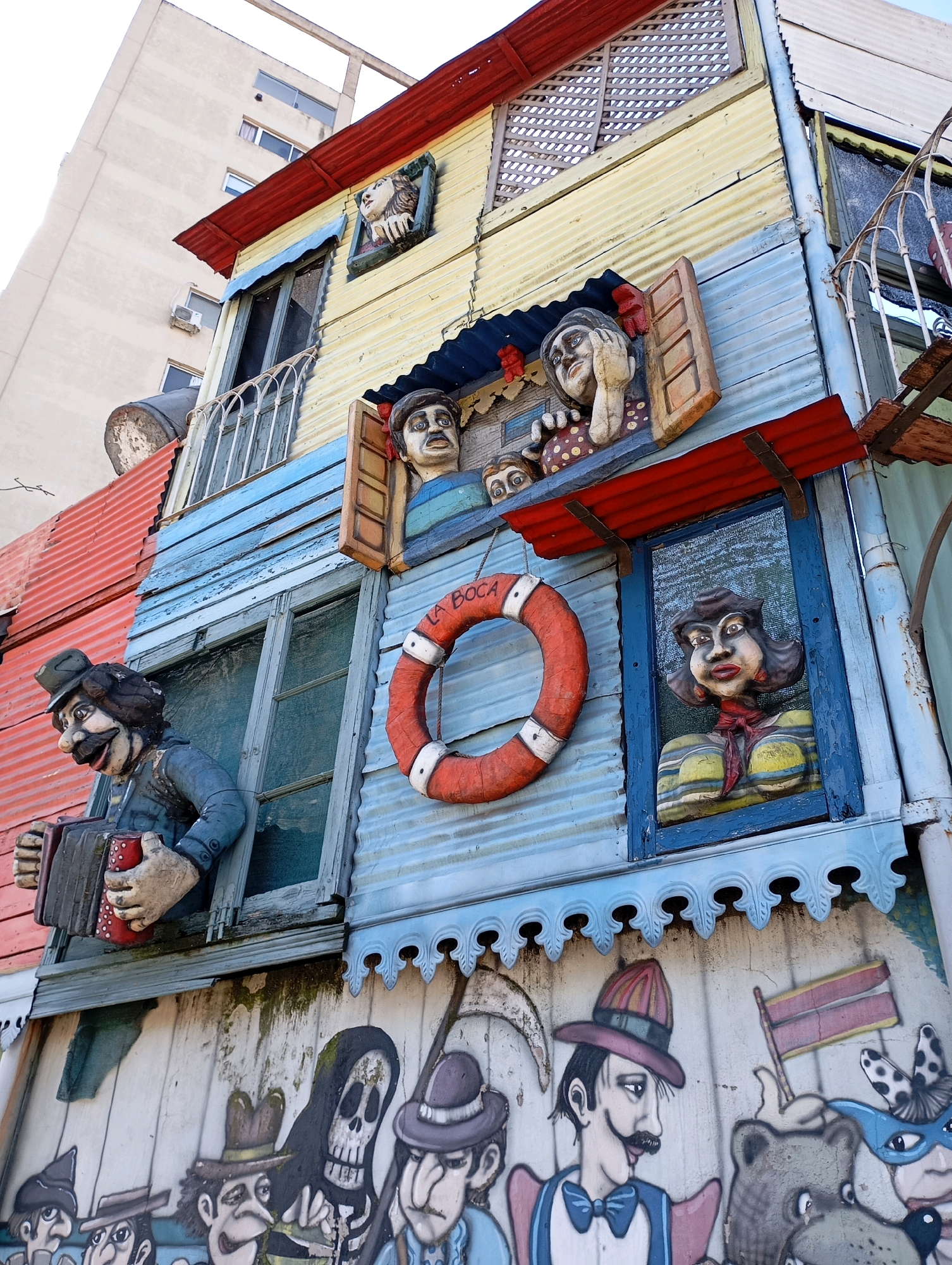 This is a low-angle, eye-level shot of the colorful, corrugated metal buildings in La Boca, Buenos Aires, Argentina. The architecture is eclectic and brightly painted, with various sculptures and murals adorning the facades. In the foreground, a sculpture of a man in a hat and blue jacket plays an accordion, peeking out from a window. To his right, a life preserver with "LA BOCA" written on it hangs from the wall. Another window features a sculpture of a woman with dark hair and red lipstick, wearing a yellow and blue striped shirt and a red scarf. Above these, other sculptures of faces are visible in windows. Below the windows, a mural depicts various characters, including a skeleton, people in hats, and animals, painted in a folk art style. The background shows a tall, modern building, contrasting with the vibrant, artistic buildings of La Boca. The sky is a pale, overcast white.