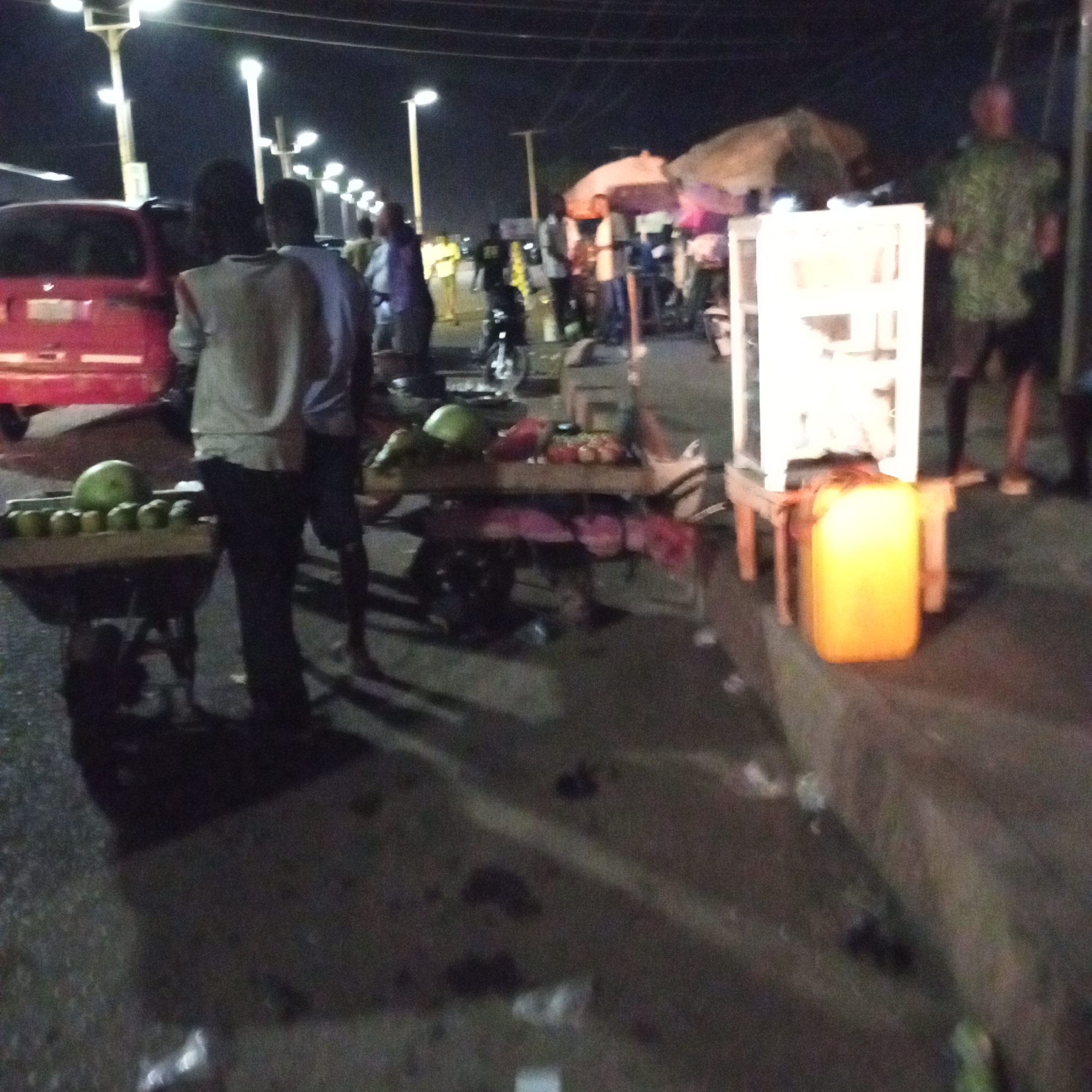 At night in Bukuru, Nigeria, street vendors are selling goods along a dimly lit roadside. The scene includes a red vehicle with its back door open, several people standing around, and carts displaying fruits and vegetables. One cart features watermelons and other produce, while another holds what appears to be a lantern or light box and a yellow jerrycan. Streetlights line the road, illuminating the scene and the activity of the vendors and customers.