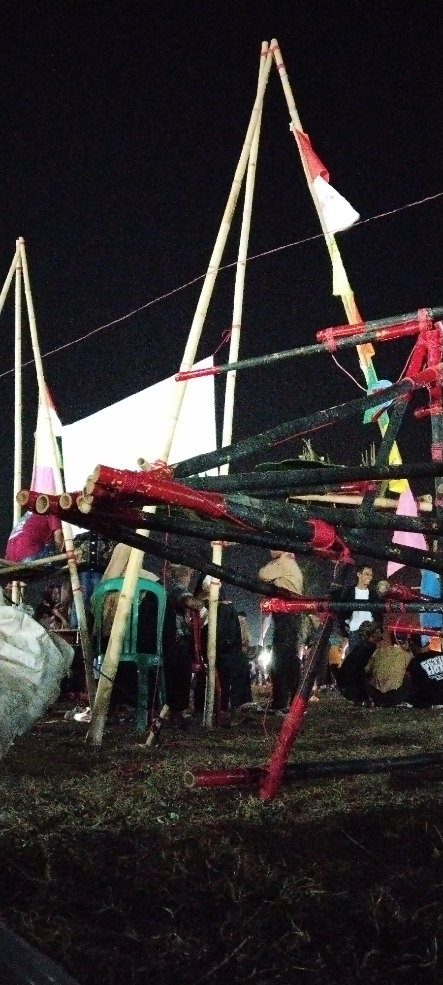 In Sido Rahayu, Indonesia, at night, a group of people are gathered around a structure made of bamboo poles and black pipes, some of which are painted red. The structure appears to be part of a temporary setup, possibly for an event or celebration. Several bamboo poles extend upwards, with a red and white flag attached to the tallest one. White fabric is stretched between some of the poles, creating a backdrop or canopy. In the foreground, a green plastic chair is visible. People are scattered around the area, some standing and some appearing to be seated or leaning. The ground is covered in dry grass and dirt. The lighting suggests artificial light sources are illuminating the scene.