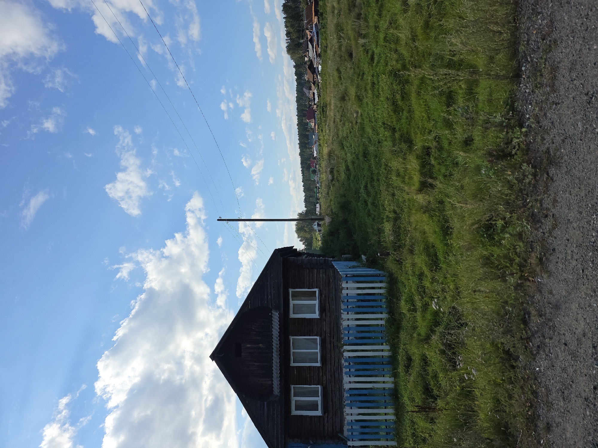 The image depicts a rural scene in Khalitova, Russia, under a bright blue sky with scattered white clouds. In the foreground, a dark wooden house with three white-framed windows and a white and blue picket fence is visible on the left. A dirt road with gravel borders the bottom of the frame. A grassy field with some tall weeds extends from the house towards the right. In the middle ground, a utility pole with electrical wires runs across the sky. Further in the distance, a cluster of houses with various colored roofs forms a small village nestled amongst trees. The overall impression is that of a peaceful, sunlit day in a quiet village.