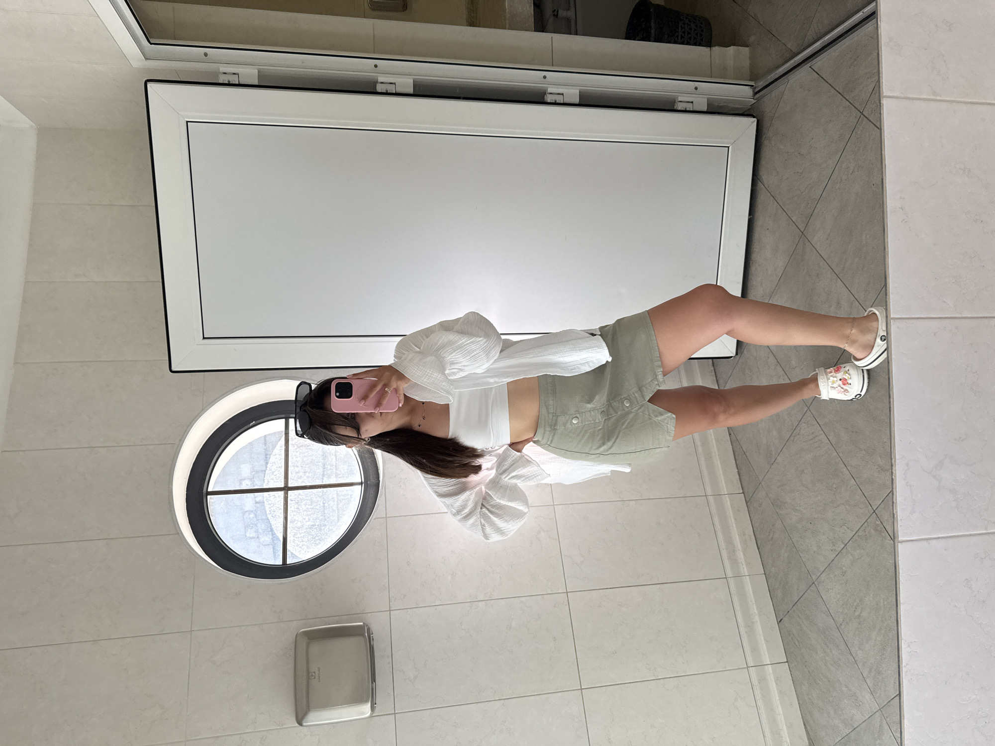 A person stands in a tiled restroom, holding a pink smartphone to take a mirror selfie. They are wearing a white bandeau top, a light green button-up skirt, and a white open-front shirt with billowy sleeves. They also have on white clogs adorned with flowers. The background includes a round window with crossbars, a silver paper towel dispenser, and a white door. The floor is tiled in a diagonal pattern of gray and white tiles. The person's reflection is visible in the large mirror.