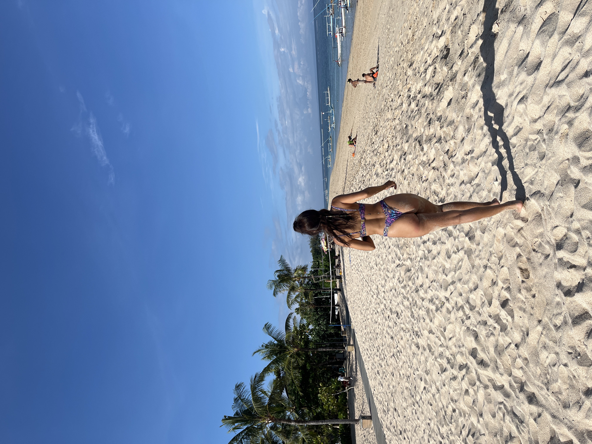 The image depicts a sunny tropical beach scene, likely in Kuta Selatan, Indonesia, as indicated by the context. The photograph is taken from behind a person, presumably a woman, who is walking on the light-colored sandy beach towards the ocean. She is wearing a patterned bikini in shades of purple and blue.
The beach itself shows ripples in the sand and distinct shadows, suggesting strong overhead sunlight, indicative of mid-day. In the mid-ground, the turquoise-blue ocean stretches out, dotted with several traditional Indonesian outrigger boats (jukung). Other individuals are visible on the beach and in the water, some appearing to be relaxing or engaging in beach activities.
To the left side of the frame, a dense line of tropical vegetation features several tall palm trees, reinforcing the island setting. The sky above is a clear, bright blue with only a few sparse, wispy clouds. No visible text is present in the image. The scene conveys a sense of warmth, relaxation, and a typical day at a beautiful tropical beach.