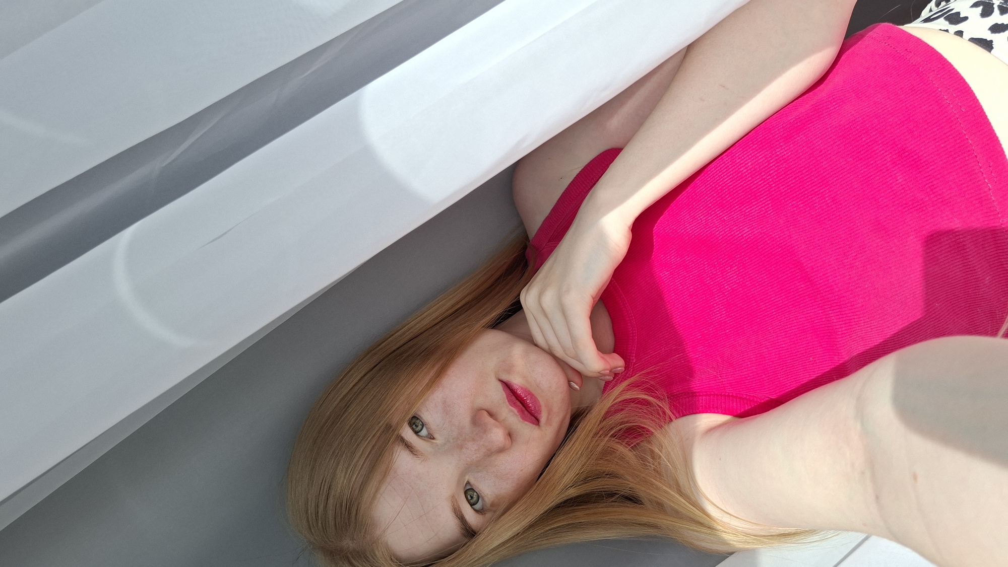 A woman is lying down. She is wearing a bright pink shirt and has long, light reddish-blonde hair. Her head is angled towards the camera, and she is looking directly at it. Her right hand is near her face. A white and gray surface is in the background. The photo appears to be taken indoors in a well-lit area. The location and city information were not provided in the image.