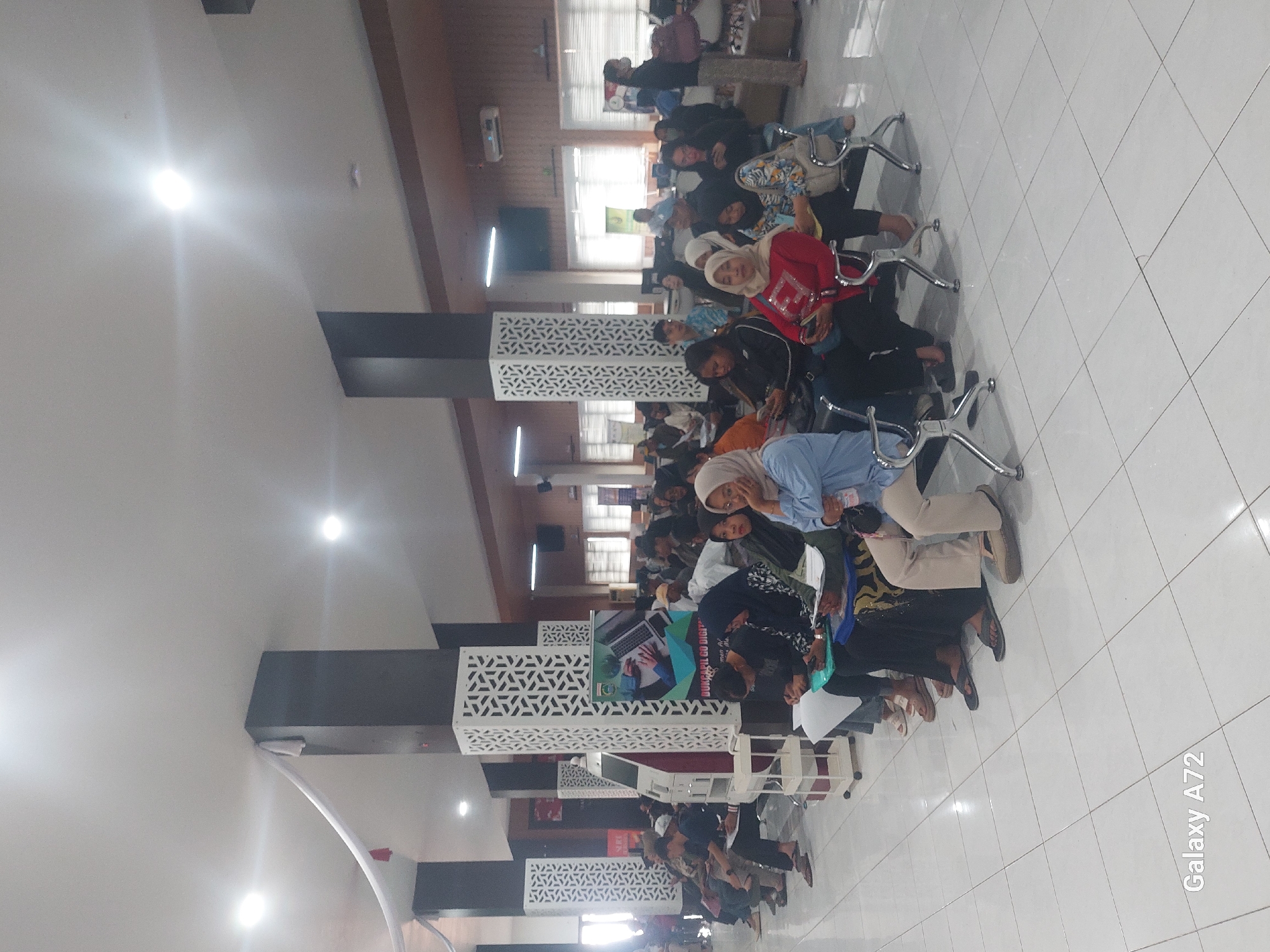 In Kedungpedaringan, Indonesia, a room with a white tiled floor and a high ceiling with recessed lighting is filled with approximately 30 to 40 people. The people are seated on a row of metal chairs, appearing to be waiting for something. Many are dressed in casual clothing, and several women are wearing headscarves. Some individuals are looking at their phones, while others appear to be engaged in conversation or looking forward. In the background, there are large structural pillars with decorative white panels and several signs, including one that reads "DUKCAPIL GO DIGITAL." A few people are standing in the background. The overall atmosphere suggests a public waiting area, possibly for government services.