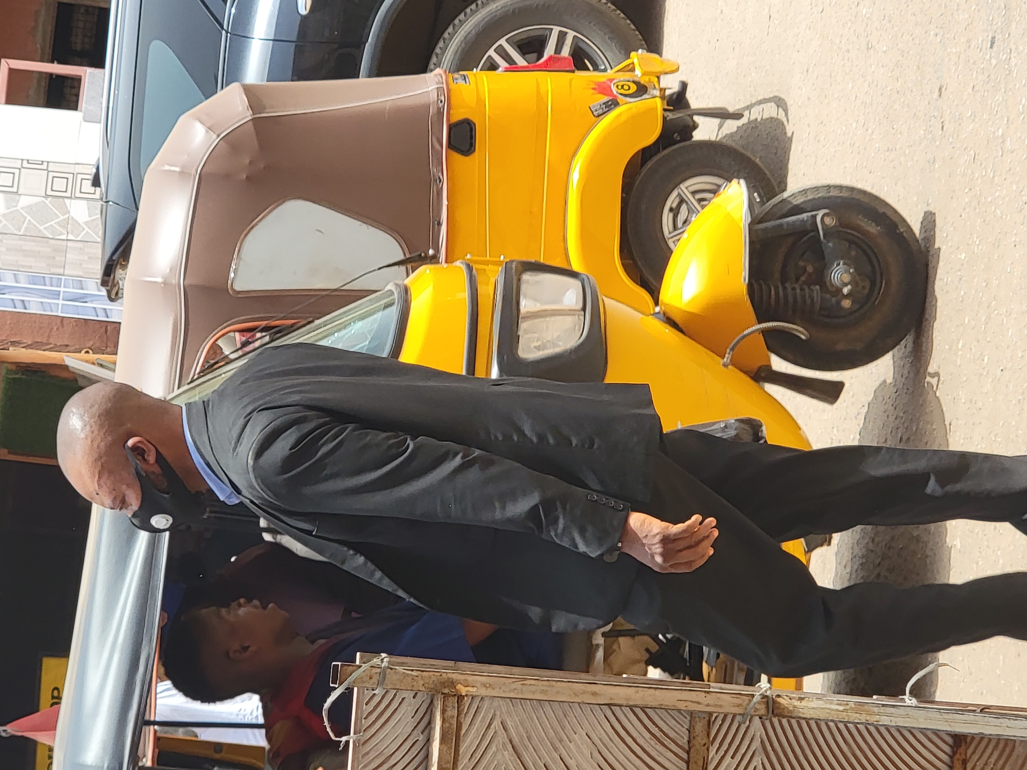 A bald man wearing a black suit and a black mask is riding in the back of a yellow auto-rickshaw. A younger man is visible in the back of the vehicle as well. The location is Maiburiji, Nigeria.