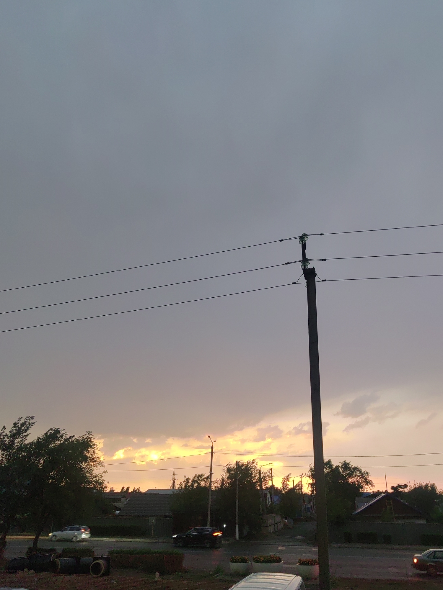 The image depicts a street scene in Ekibastuz, Kazakhstan, during sunset. A concrete utility pole with multiple power lines stretches vertically through the right side of the frame. The sky above is overcast with a gradient of gray to pale orange and yellow hues, indicative of the setting sun. On the left side of the image, a dense tree with dark foliage stands prominently. In the middle ground, houses with dark roofs are visible, interspersed with trees and a street. Several vehicles are present: a white SUV drives on the left side of the street, and a dark sedan is visible on the right. The foreground shows a street with what appear to be concrete pipes and some potted plants. The overall atmosphere is muted, with the warm glow of the sunset creating a dramatic contrast against the gray sky.