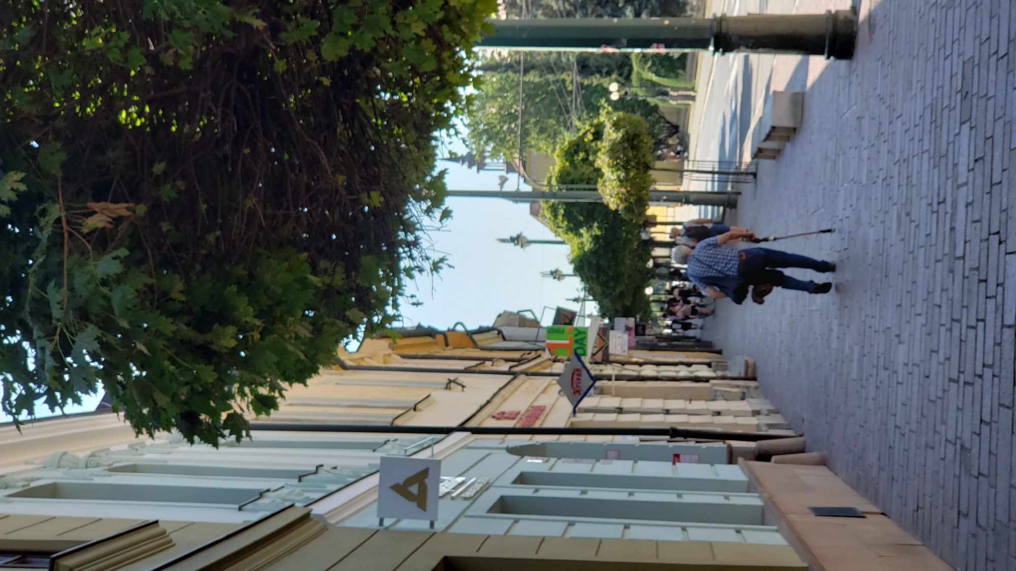 A street scene in Prešov, Slovakia shows a man walking on a brick sidewalk, using a cane. He is wearing a blue and white checkered shirt and blue jeans and carrying a bag over his shoulder. The sidewalk is lined with trees and shops. In the background, a road is visible with traffic and more trees. The architecture suggests a European city with old buildings.