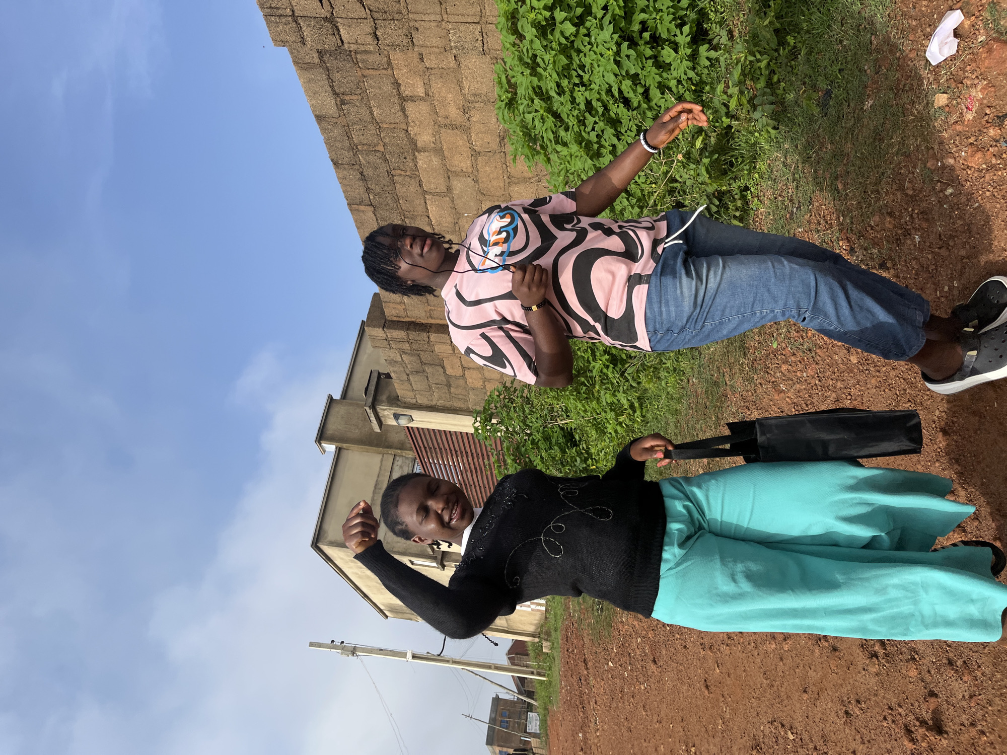 Two young women are walking side-by-side on a dirt path in Jos, Nigeria, with a building and a brick wall in the background. The woman on the left, wearing a black sweater and wide teal pants, is smiling and has her left arm raised in a fist-pump gesture. She is carrying a black tote bag in her right hand. The woman on the right, wearing a pink and black patterned t-shirt and blue jeans, is looking towards the camera and holding white earbuds. Lush green vegetation is visible behind them, and the sky is partly cloudy with a bright blue hue. The ground is covered in reddish-brown dirt, typical of the area. A white piece of paper is on the ground in the lower right corner of the frame.