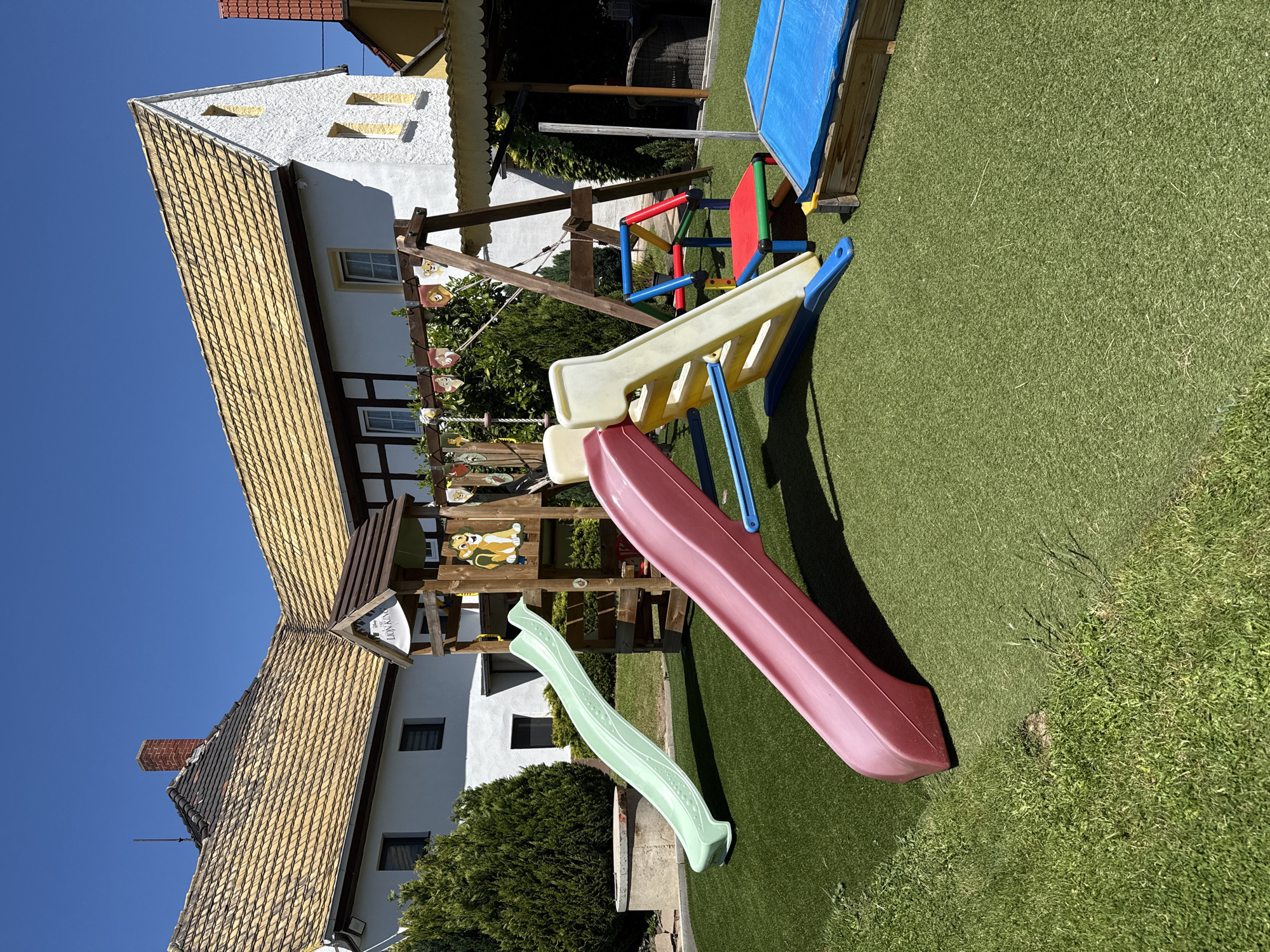 The image shows a backyard playground with a slide, swing set, and a covered sandbox. The playground is situated on artificial turf. In the background is a traditional German building with a tiled roof, half-timbered construction, and white stucco walls. The location is likely Zürchau, Germany. No people are visible in the image.