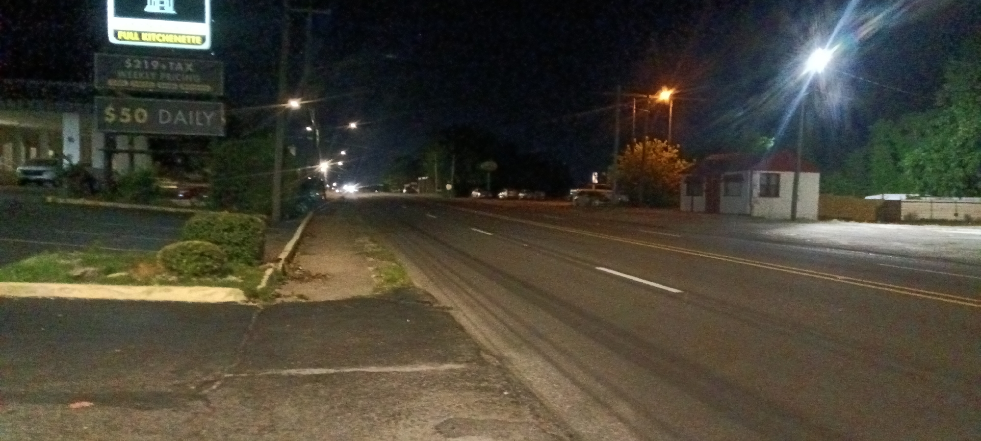 The image depicts an urban street scene at night. The location is identifiable by the business sign to the left, which indicates "Full Kitchenette," "$219+TAX Weekly Pricing," and "$50 DAILY." The street is a paved road with streetlights illuminating the area. The road has two lanes on each side, separated by a white line. Visible tire tracks are present in the lanes. On the left side of the road is a sidewalk with foliage and a building further back. On the right side is a small white structure with a red roof, set back from the road. Further down the road, headlights from distant vehicles can be seen. The environment suggests a commercial or residential area in Fort Smith, Arkansas, United States.
