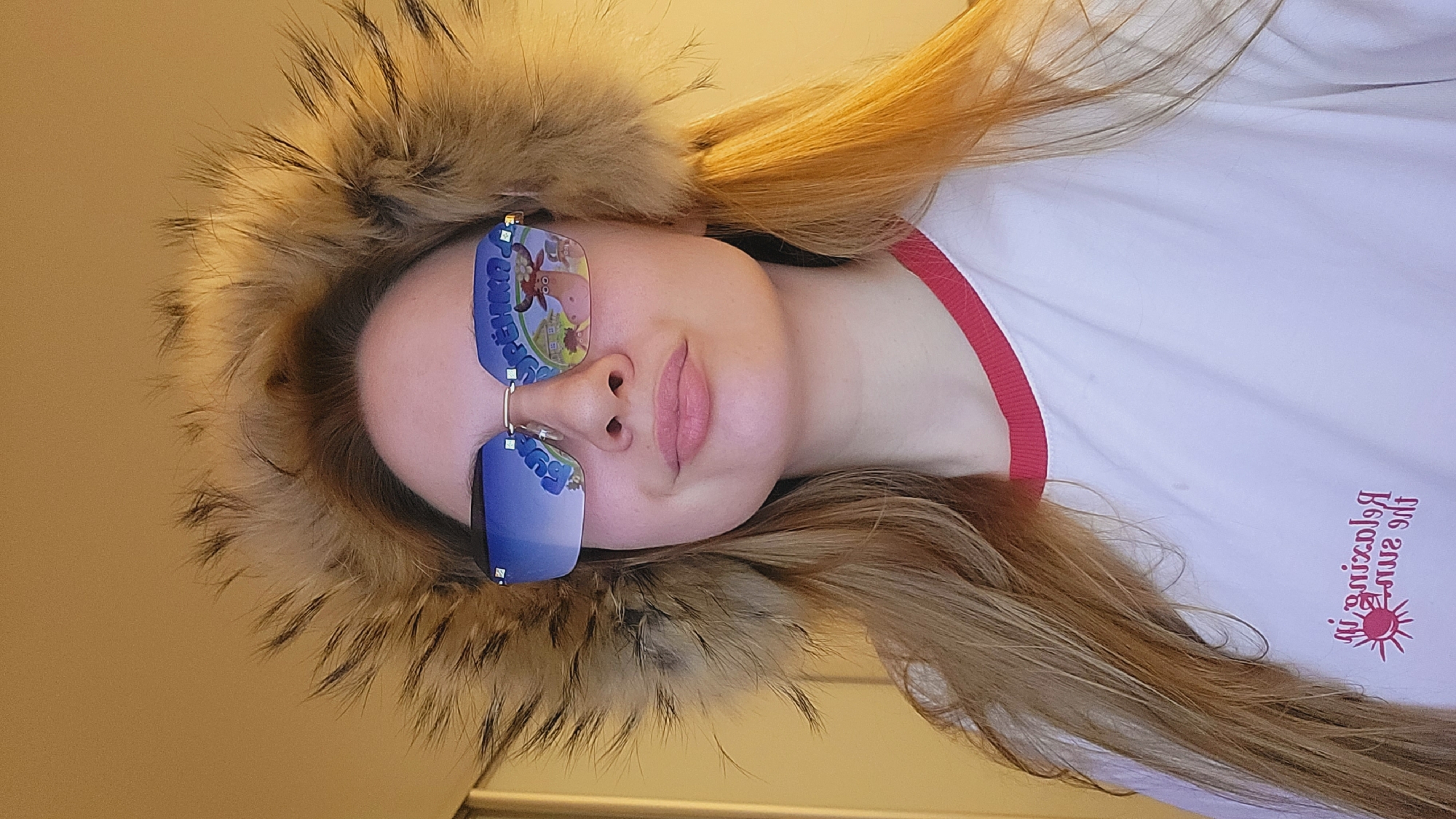 A young woman wearing a fur-trimmed hood and sunglasses stands indoors. The reflection in her sunglasses shows an image of cartoon characters. She wears a white shirt with red trim. Text on her shirt reads "Relaxing at the Sun". There is no information indicating that this image was taken in Chaikovsky, Russia.