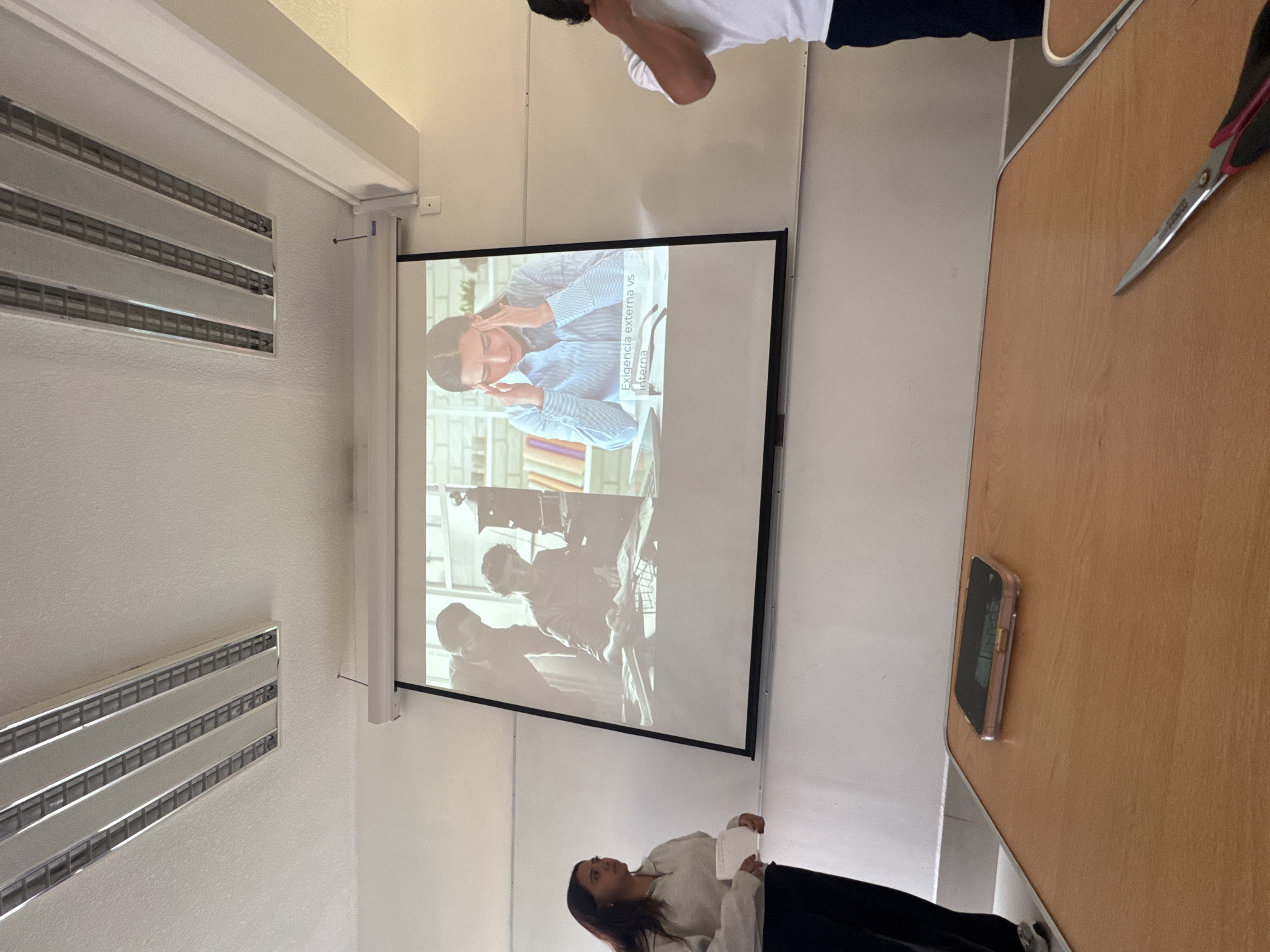 The image shows a classroom-like setting in San Andrés Cholula, Mexico. A projector screen displays two images: a woman holding her head, alongside the text "Exigencia externa vs interna," and a black and white image of a man holding a film camera. The environment includes a wall with air conditioning vents, a table, and a phone on the table. People visible in the image include a person in a white shirt and navy pants, and a person wearing a beige top and black pants. Scissors are placed on the table.