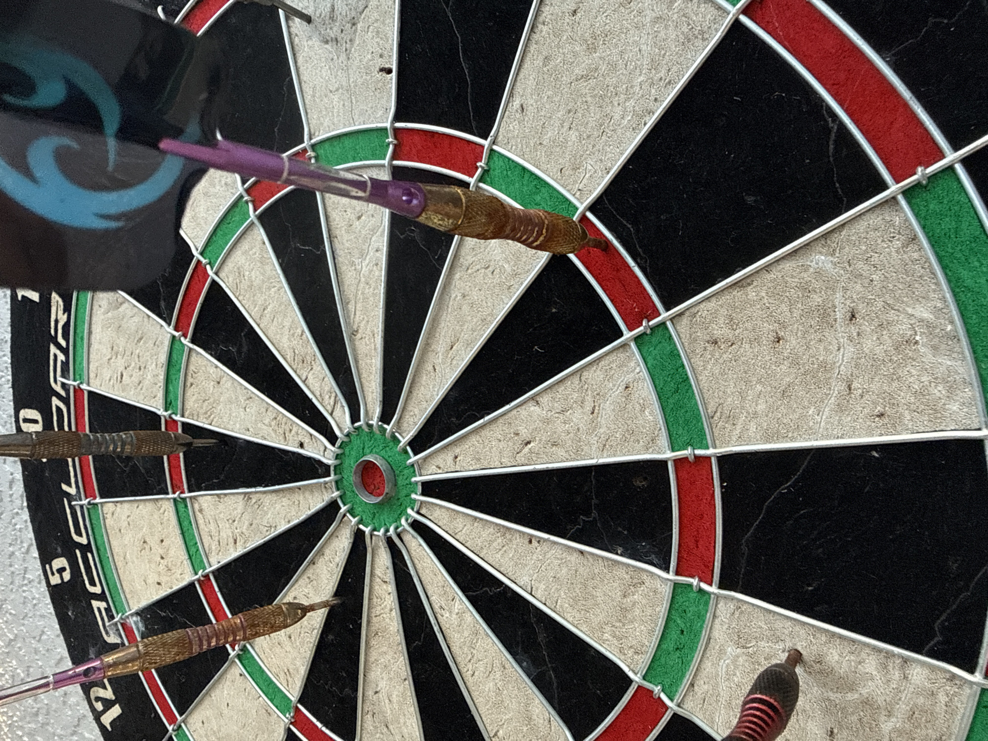 In Trinity, United States of America, a close-up, high-angle shot captures a dartboard with several darts embedded. The dartboard is circular, divided into segments by thin metal wires. The segments alternate between black and beige, with outer rings of red and green. The center of the board features a small red bullseye.
Five darts are visible. One dart, with a purple fletching and a gold barrel, is positioned vertically, its tip inserted into the green ring between two black segments. Another dart, predominantly gold with thin red stripes and a pink fletching, is stuck in a beige segment near the top left of the board. A third dart, dark in color with a red and black striped fletching, is lodged in a beige segment on the lower left. Two more darts are present; one, with a dark fletching and a gold barrel, is stuck in the upper right portion of the board, and another dart with a purple fletching is visible with its tip just above the board on the right side. The surface of the dartboard appears worn.