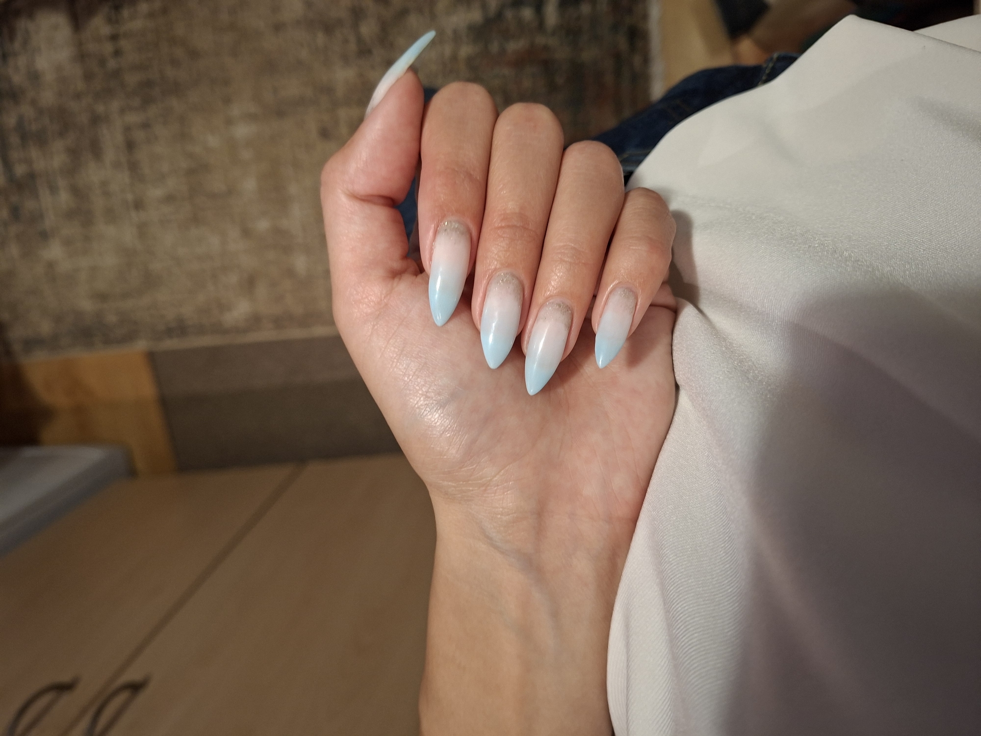 A person's hand with light blue and white ombre stiletto-shaped nails rests on a white fabric. The nails have a subtle glittery detail near the cuticle. The background is blurred, showing wooden furniture and a textured rug. The lighting is artificial, casting a warm glow on the skin. The location is unspecified.