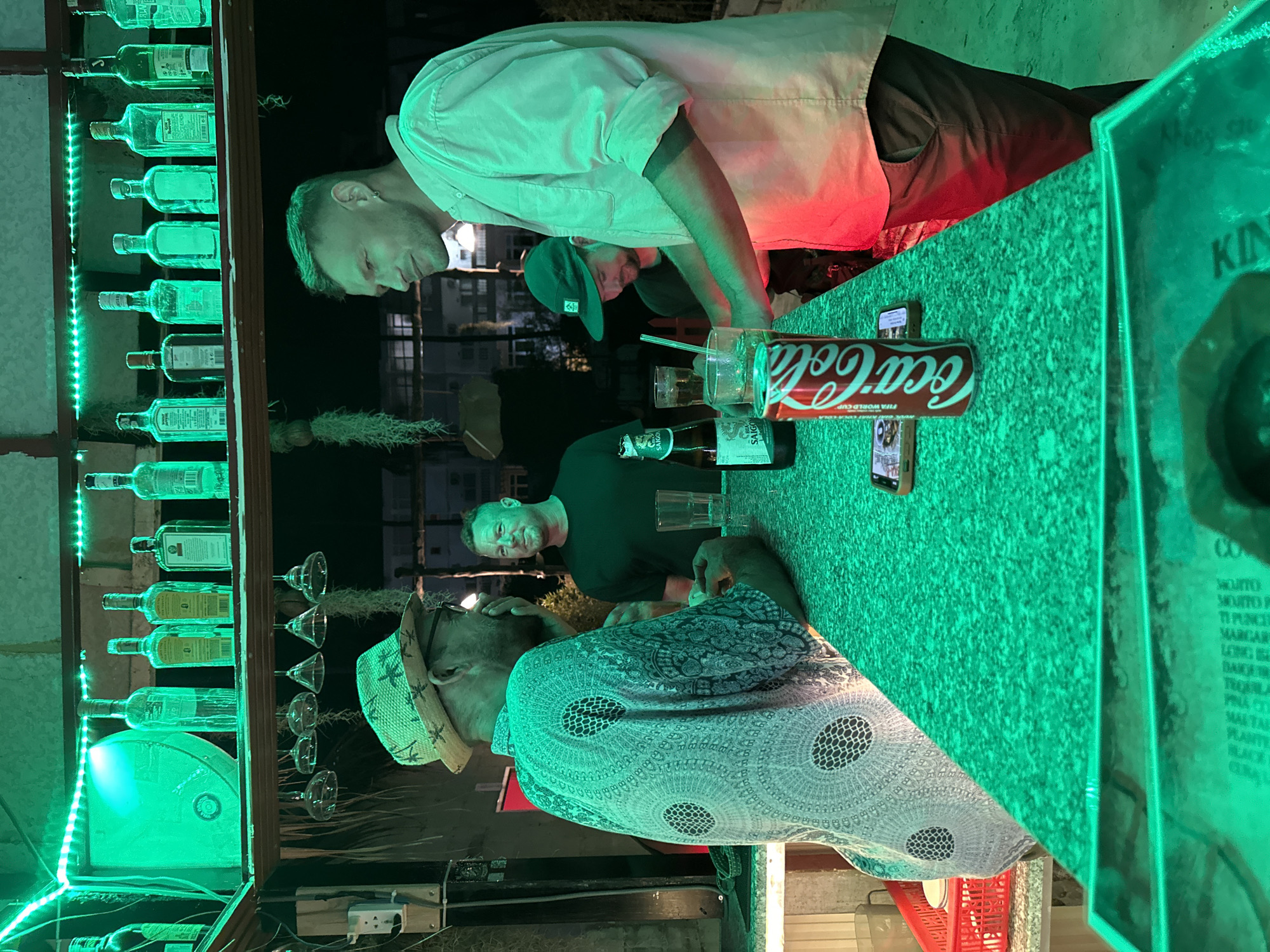The image shows a nighttime scene at a bar in Mũi Né, Vietnam. The focus is on the interaction between the bartender and several patrons.
A light-skinned bartender in a white shirt leans over the green-lit bar, attending to customers. Among the customers are several light-skinned men, one wearing a green cap and another in a black shirt. One customer is wearing a straw hat and a patterned shirt. A Coca-Cola can and beer bottles are on the bar.
The bar is decorated with green LED lights and a shelf displaying liquor bottles. A menu board with text "KIN" and a list of cocktails is visible. The environment outside the bar includes a view of a building.