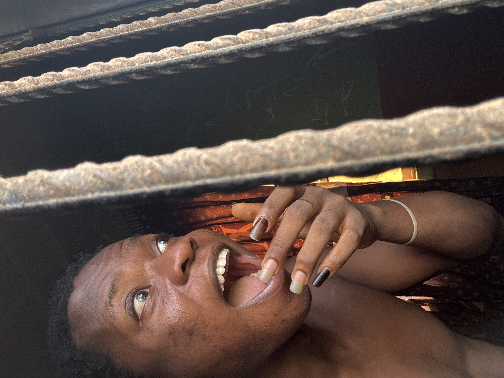 A person is lying on their back, seemingly under a grate. They are looking up with their mouth open, and one hand is raised near their face. The fingernails are painted in a varied style, including a mix of brown, bare, and black. The background is obscured by the grate and appears to be a mix of dark and light areas. A bracelet is seen on the wrist. The location is unknown.