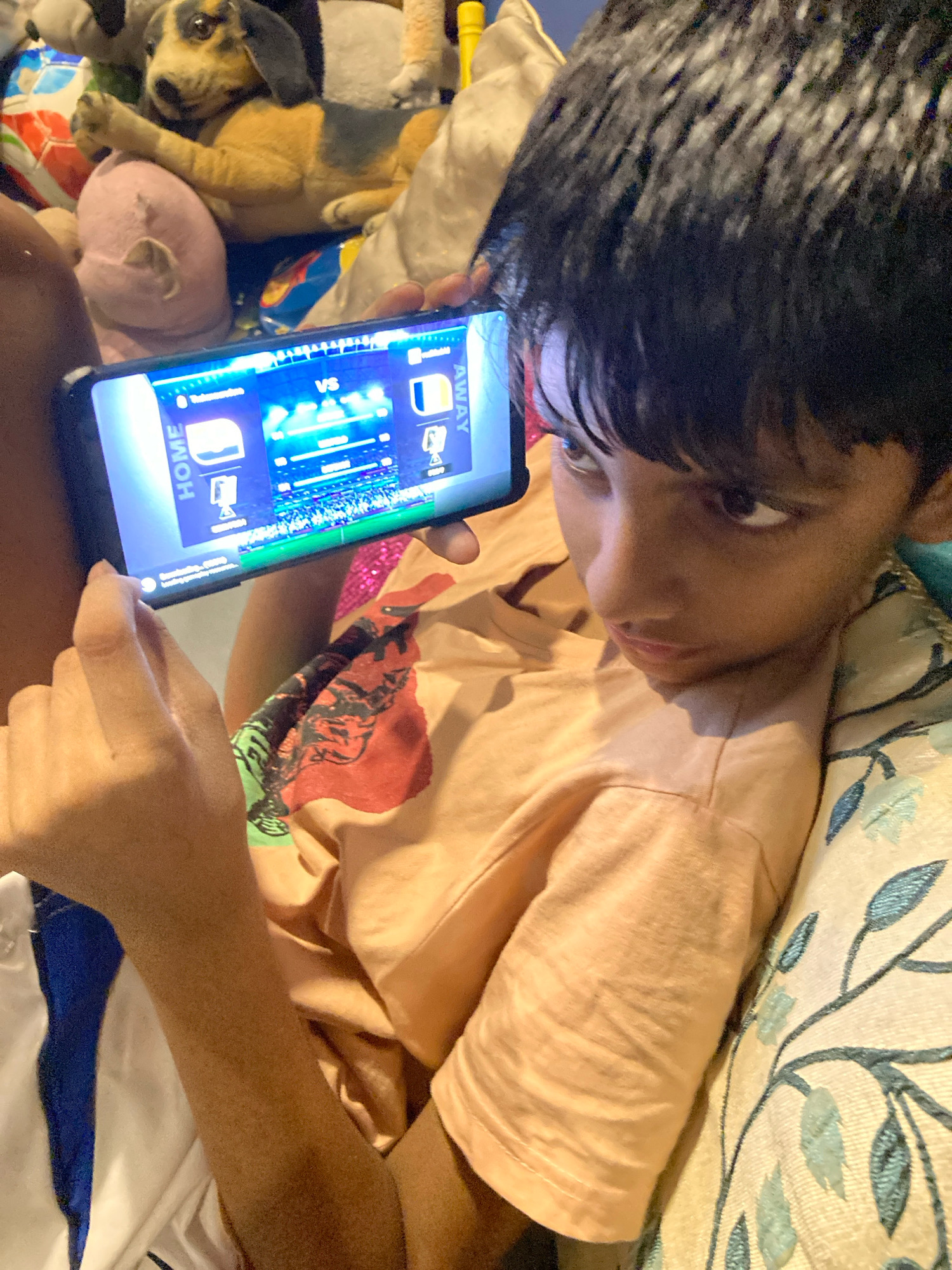 A young boy, likely of South Asian descent, with dark hair and brown eyes, is shown from the chest up, holding a smartphone horizontally in his hands. He is wearing a light orange t-shirt with a faded red and black graphic print. The boy is lying down on a sofa with a patterned cushion. The phone displays a sports game interface with "HOME" and "AWAY" labels, suggesting a match is in progress. The background is cluttered with stuffed toys, including a dog and a pink animal, some of which are out of focus. The lighting suggests an indoor setting, possibly a living room. There is no clear indication of the location being specifically in Kolkata, India.