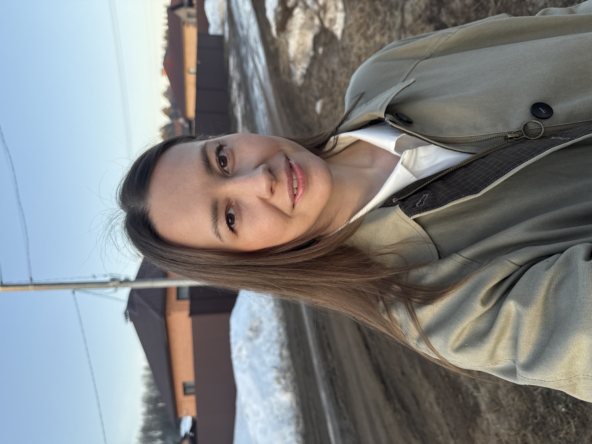 A woman with long brown hair is smiling at the camera. She is wearing a khaki jacket over a white shirt. The background includes a road with melting snow, houses, and a blue sky. The location is likely Chaikovsky, Russia. There are power lines visible against the sky.