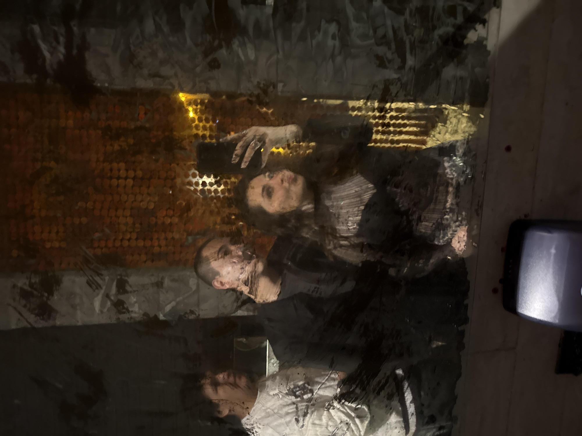 The image shows three people reflected in a dirty, possibly glass, surface. Two individuals are closer to the camera; a woman holding a smartphone, seemingly taking a picture, and a man slightly in front of her. A third person is reflected at the bottom of the frame. The background has a textured, patterned appearance that is obscured by dirt or smudges. A dispenser-like object is partially visible on the right side of the frame. There are no readily identifiable architectural features to confirm the location in San Andrés Cholula, Mexico.
