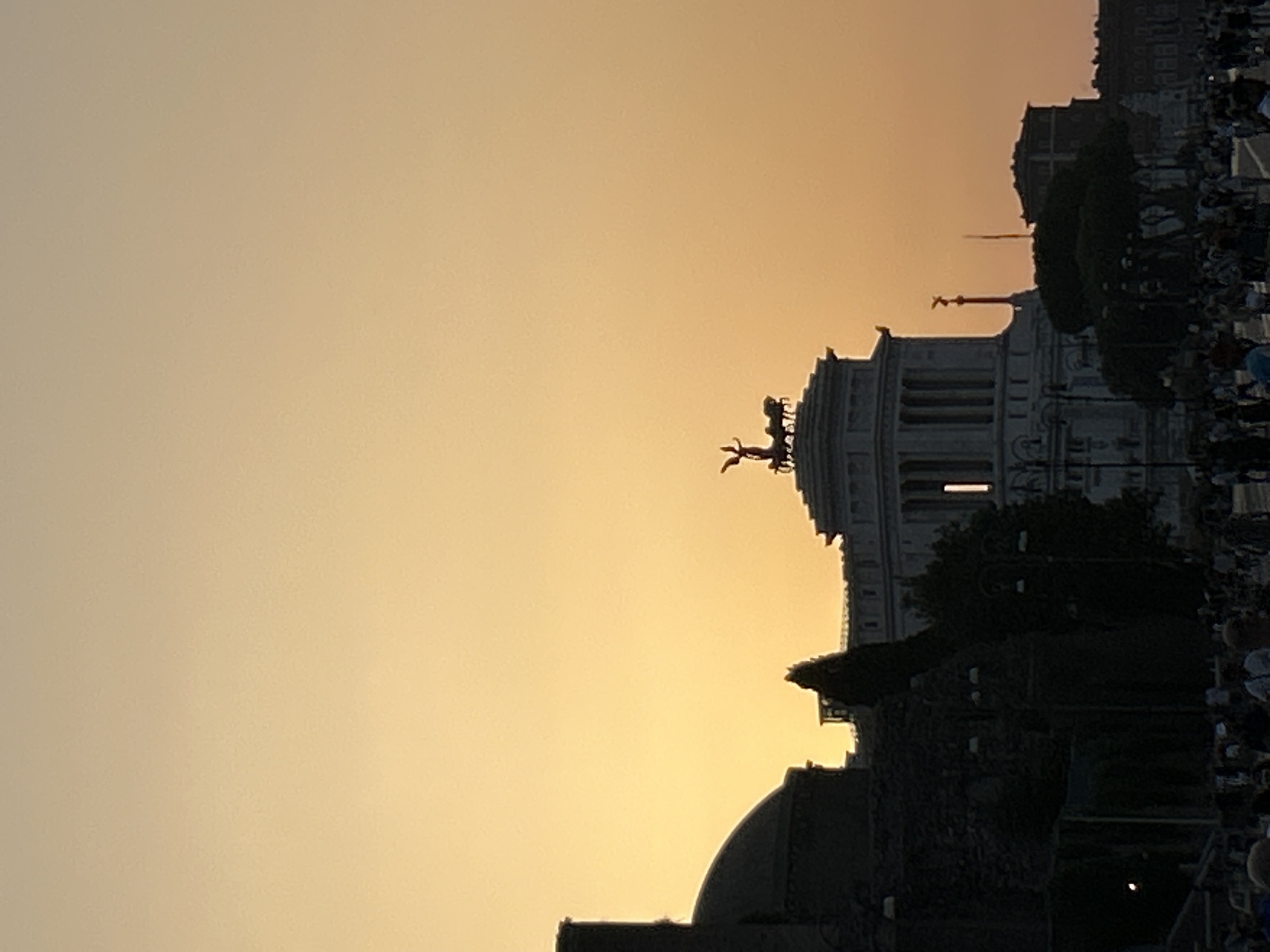 The image depicts a sunset over Rome, Italy. In the foreground, a crowd of people is visible, suggesting a public space or event. The silhouette of a large, imposing building, possibly a monument or historical structure, dominates the mid-ground. On top of this building, a sculpture of a chariot pulled by horses is clearly discernible against the orange sky. Further in the background, other buildings and trees are silhouetted. The sky is a gradient of warm colors, transitioning from a pale yellow at the top to a deeper orange towards the horizon, indicative of a sunset. The overall scene captures a moment of dusk in the Italian capital, with the iconic architecture and the atmospheric lighting creating a picturesque view.