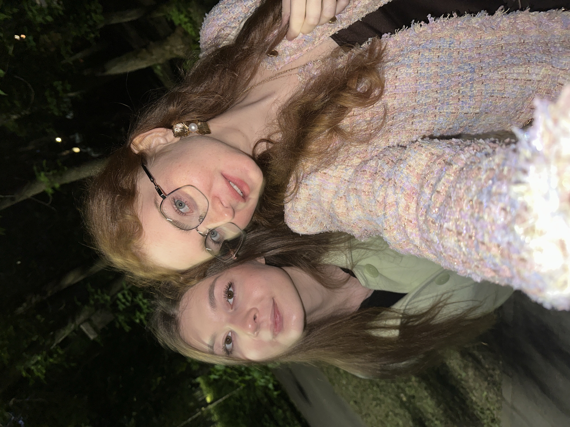 The image is a nighttime selfie taken outdoors in Kazan, Russia, featuring two young adult women.
The woman on the left has long, straight, dark brown hair parted slightly off-center and falling over her shoulders. She has brown eyes, a straight nose, and her lips are slightly parted in a gentle smile. She wears a light, possibly mint green, collared jacket with two visible round, light green buttons on the front closure, over a dark undershirt. Her skin tone is light.
The woman on the right, positioned in the foreground and closer to the camera, has long, wavy, auburn hair. She has blue eyes, light freckles on her nose and cheeks, and her lips are subtly colored, appearing to have a light gloss. She wears eyeglasses with thin, angular metal frames. Her left ear (viewer's right) displays a prominent gold-toned earring shaped like a fan or leaf, accented with a white pearl in the center. She is wearing a textured, woven jacket made of pastel-colored threads including light pink, blue, purple, and white, giving it a tweed-like appearance with subtle shimmer and frayed edges along the opening. Small, round gold-toned buttons are visible on the jacket. A delicate gold-toned chain necklace is visible over her dark brown top. Her left hand (viewer's right) is subtly holding the front edge of her jacket. Both women are looking directly at the camera with soft smiles.
The background is dark, showing the blurred silhouettes of trees, indicating a natural outdoor setting such as a park or garden. Faint, out-of-focus lights are visible among the trees in the distance. A paved surface is partially visible in the lower left of the frame. The lighting is focused on the subjects, suggesting the use of a flash or a nearby light source.