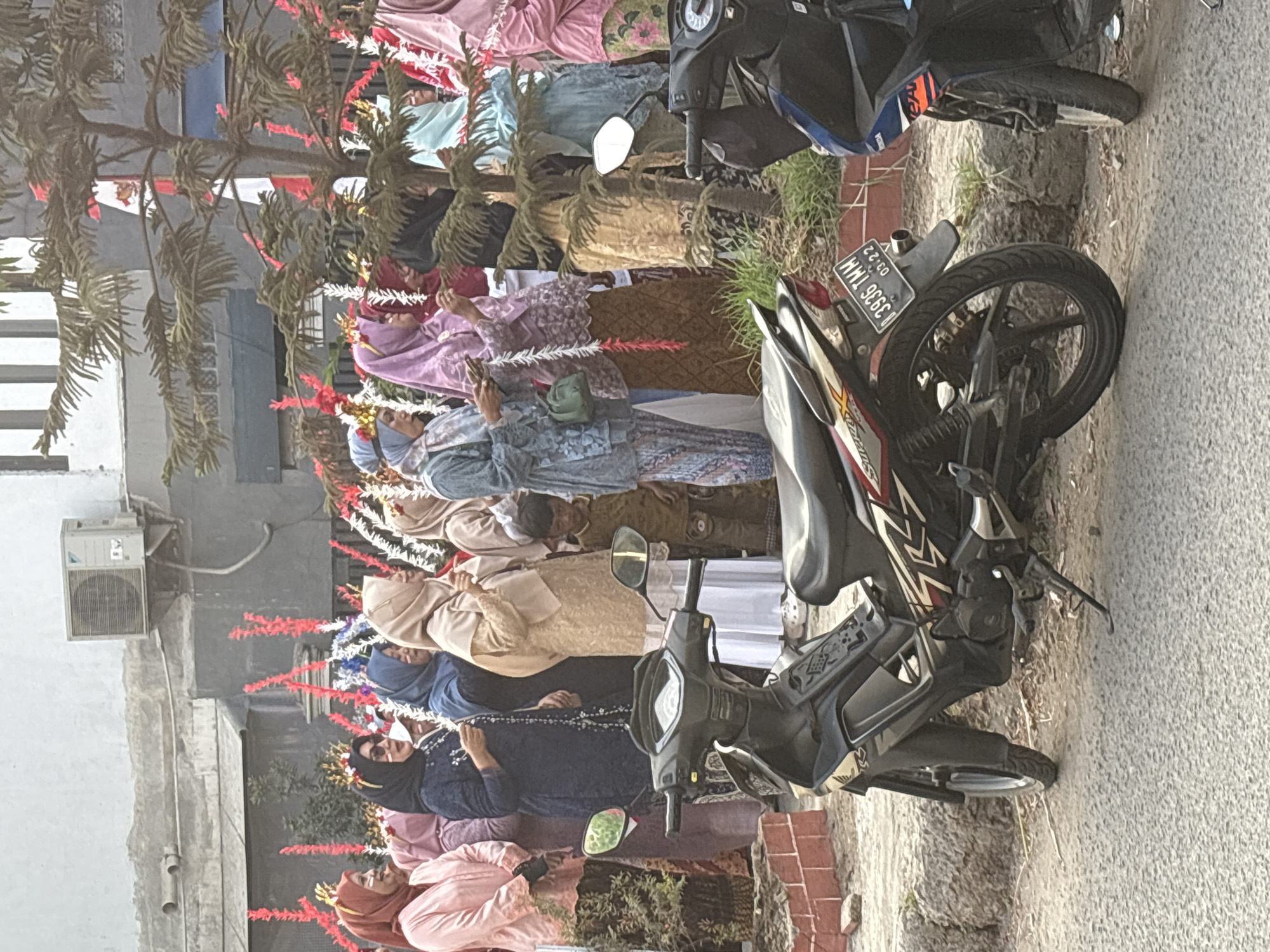 A group of Muslim women, some wearing elaborate headwear and festive decorations, stand in a line outdoors. The women are dressed in various colorful hijabs and traditional attire. A young boy in a brown shirt stands behind them. In the foreground, two motorcycles are parked on a paved surface. One motorcycle is black and white, and the other is blue and black. The environment appears to be a street or sidewalk in a residential area. A building with an air conditioning unit is visible in the background. The scene is likely in Telagamurni, Indonesia, suggested by the license plate on the black and white motorcycle which reads "B 3936 TMM" and the format of the date "03.22".