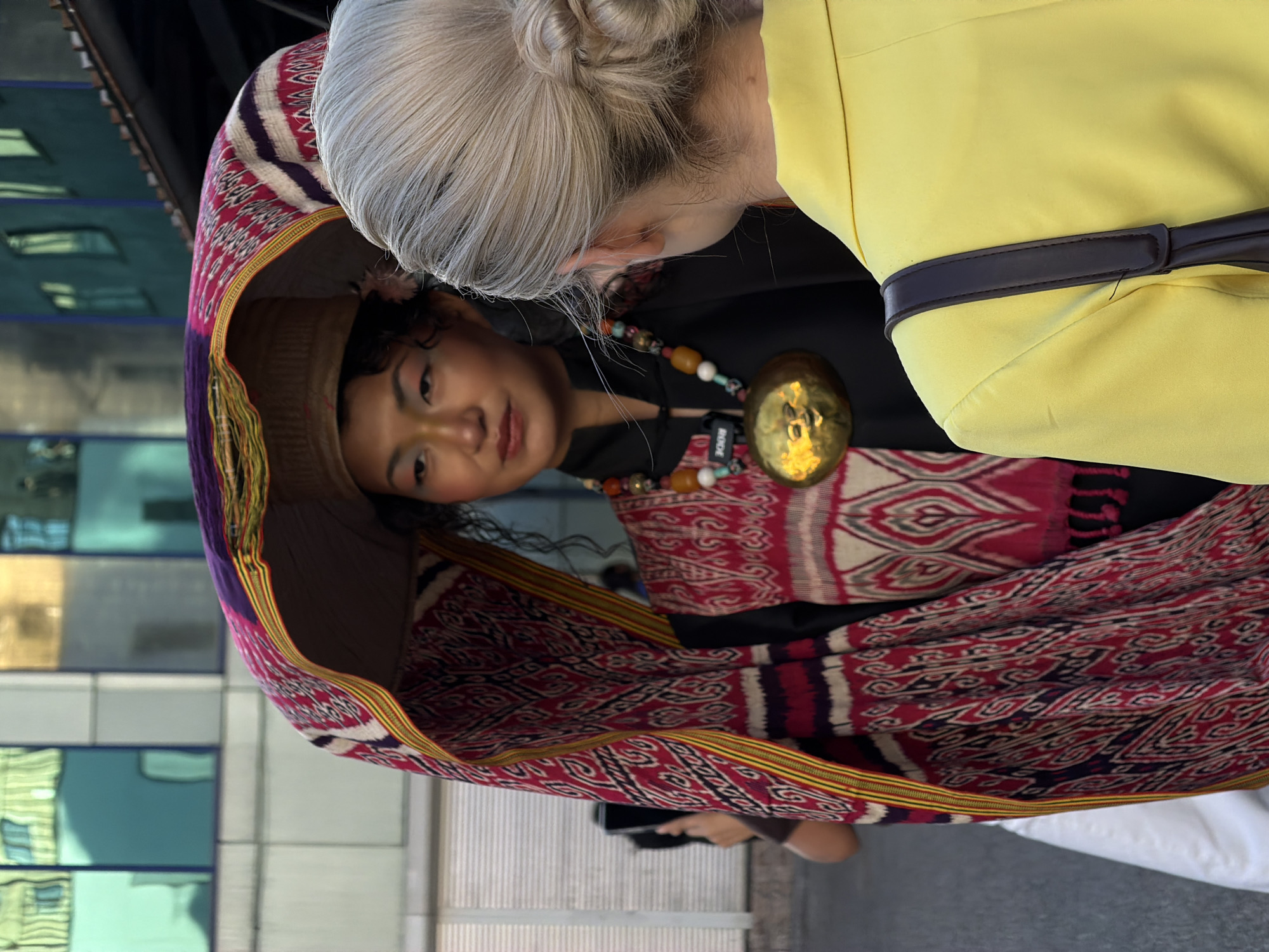 A horizontal shot captures a woman in traditional Kyrgyz attire, gazing up and to the left at another woman with blonde hair styled in a bun. The blonde woman, wearing a yellow blazer with a dark brown belt, is positioned with her back to the camera, her attention focused on the Kyrgyz woman.
The Kyrgyz woman is draped in a vividly patterned red and purple cloth with ornate detailing, which extends into a hood that frames her face. She is also wearing a black top and a necklace with amber beads and a large golden pendant. A Rode microphone is clipped to her clothing.
The photograph appears to have been taken in Bishkek City, Kyrgyzstan. In the blurred background, a modern building with reflective glass panels indicates an urban environment. The street or walkway is visible in the foreground, suggesting an outdoor setting. The image's overall composition captures an interaction between two individuals against an urban backdrop, highlighting the Kyrgyz woman's traditional attire and the contrast with the modern surroundings.